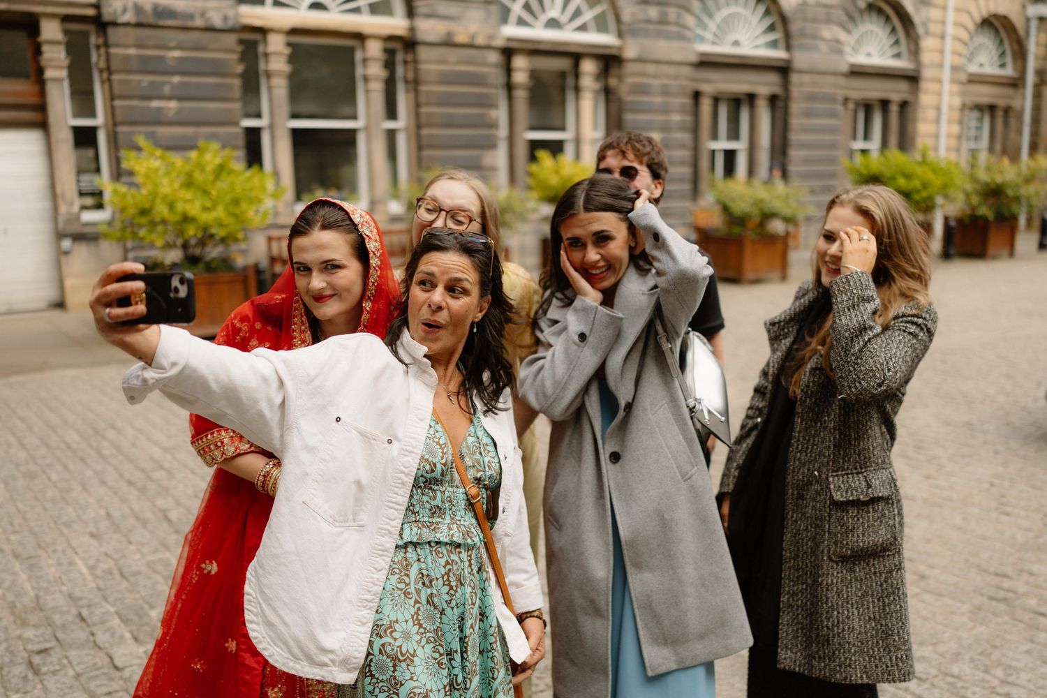 Bride in red dupatta takes selfie with group of smiling women outside historic building.