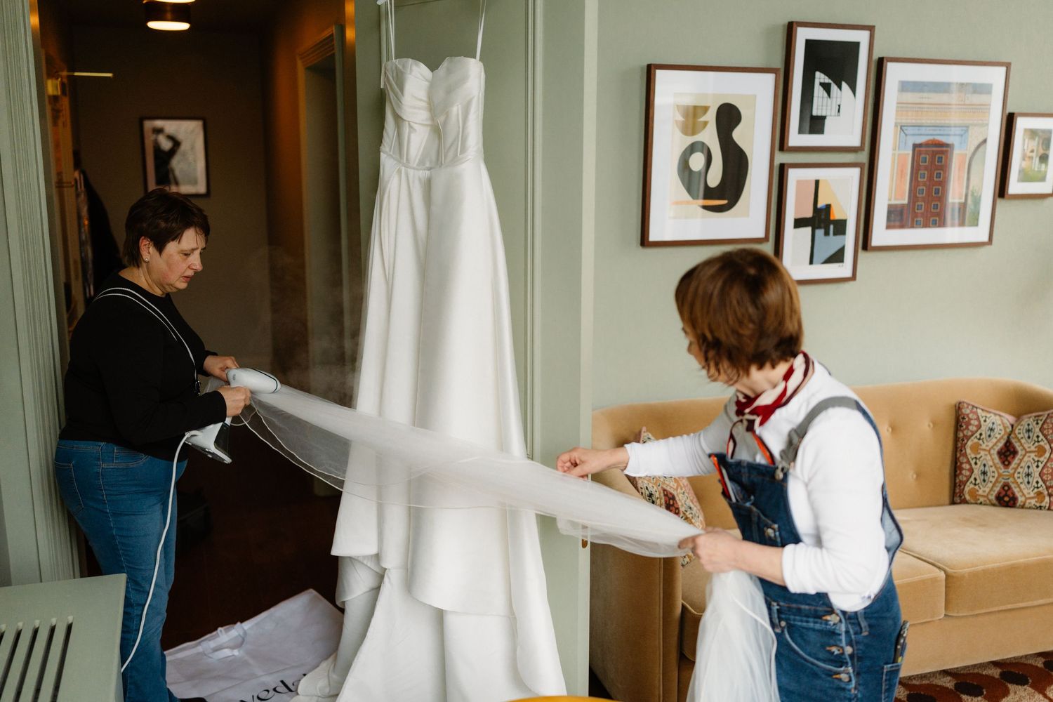 Two women steaming and preparing a white wedding dress hanging in a elegant room with gallery wall.