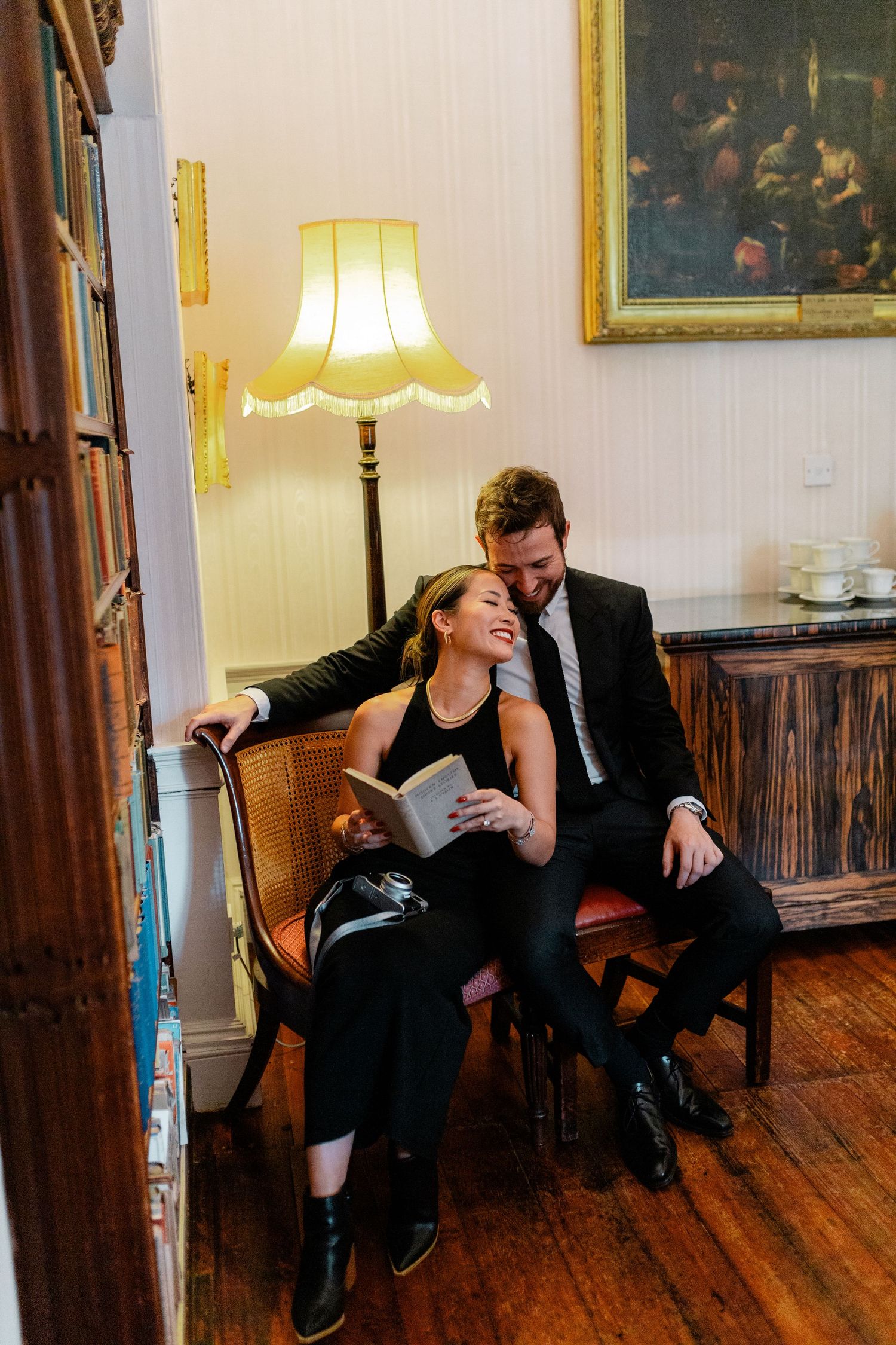 A couple shares a romantic moment reading a book together in a cozy, lamp-lit library room.