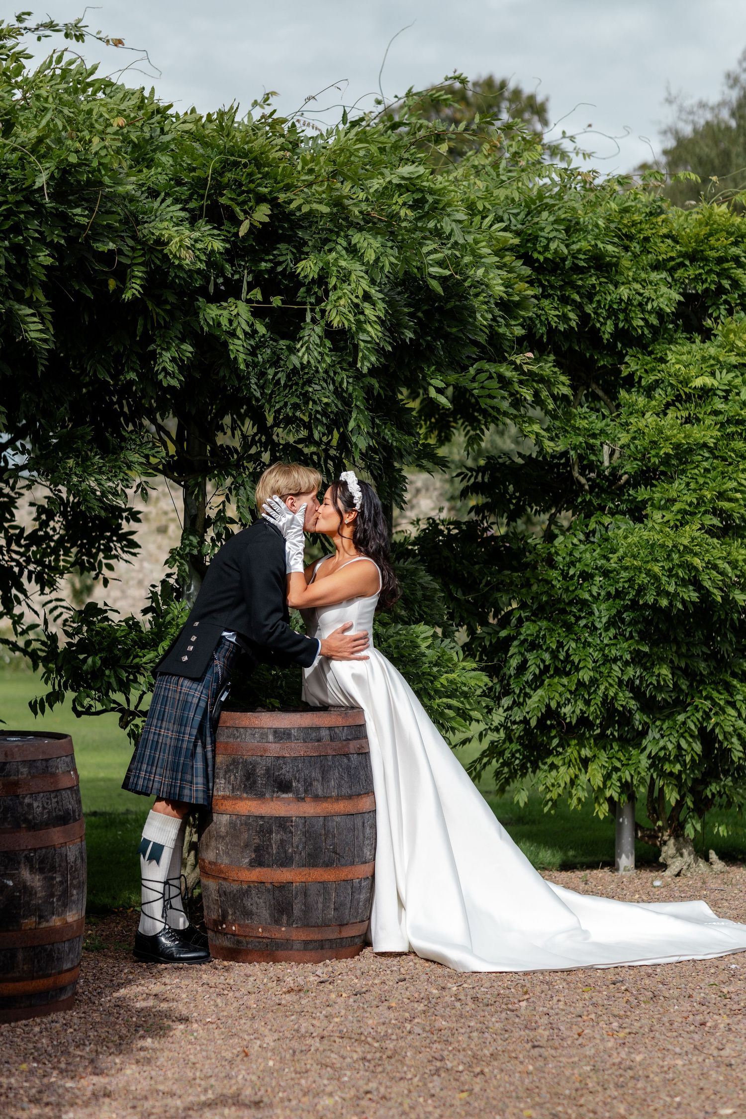Wedding couple posing by rustic wooden barrels in garden setting with flowing white dress.