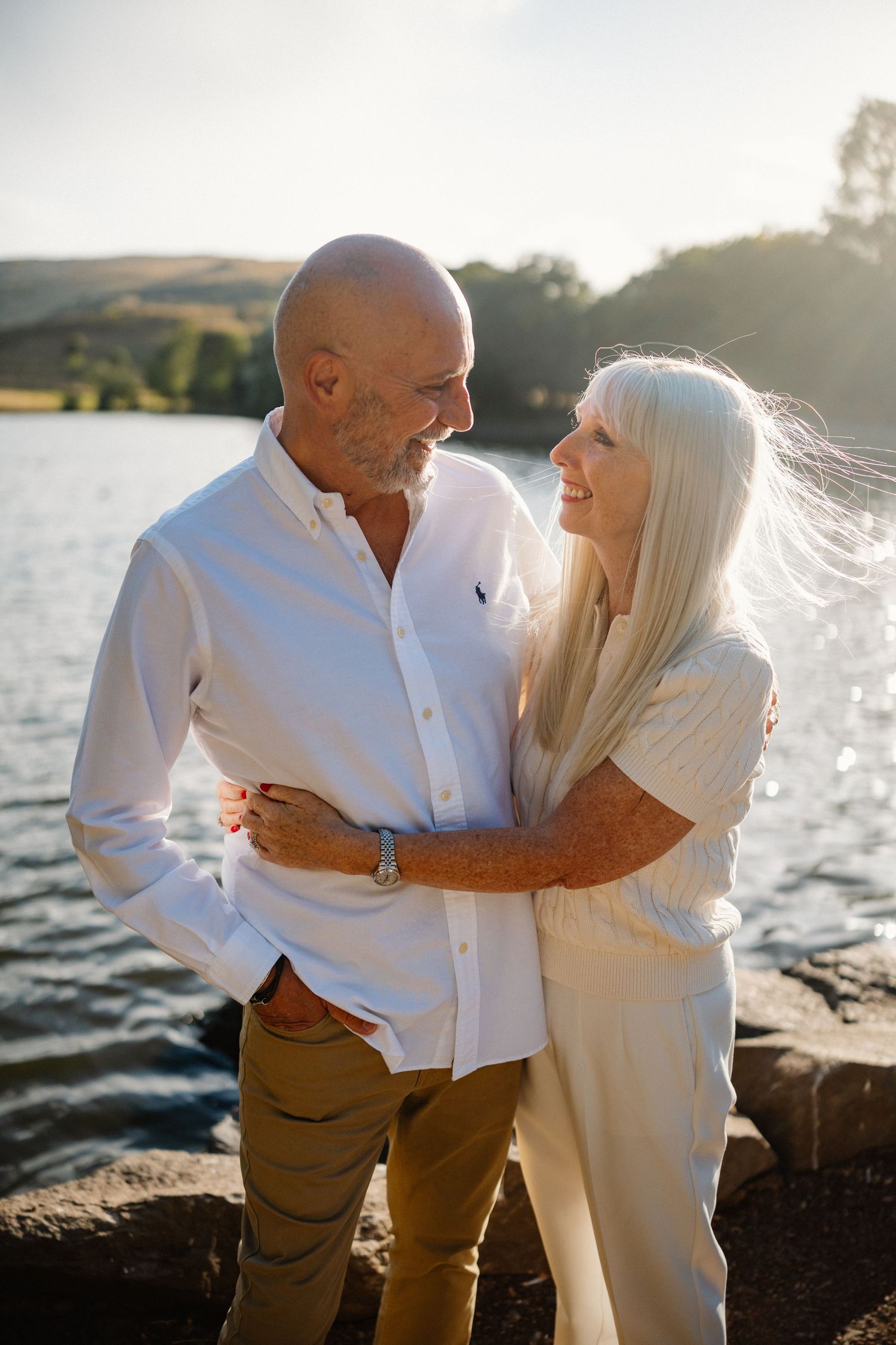 Couple in white and tan clothing sharing a romantic moment by a lake at sunset.