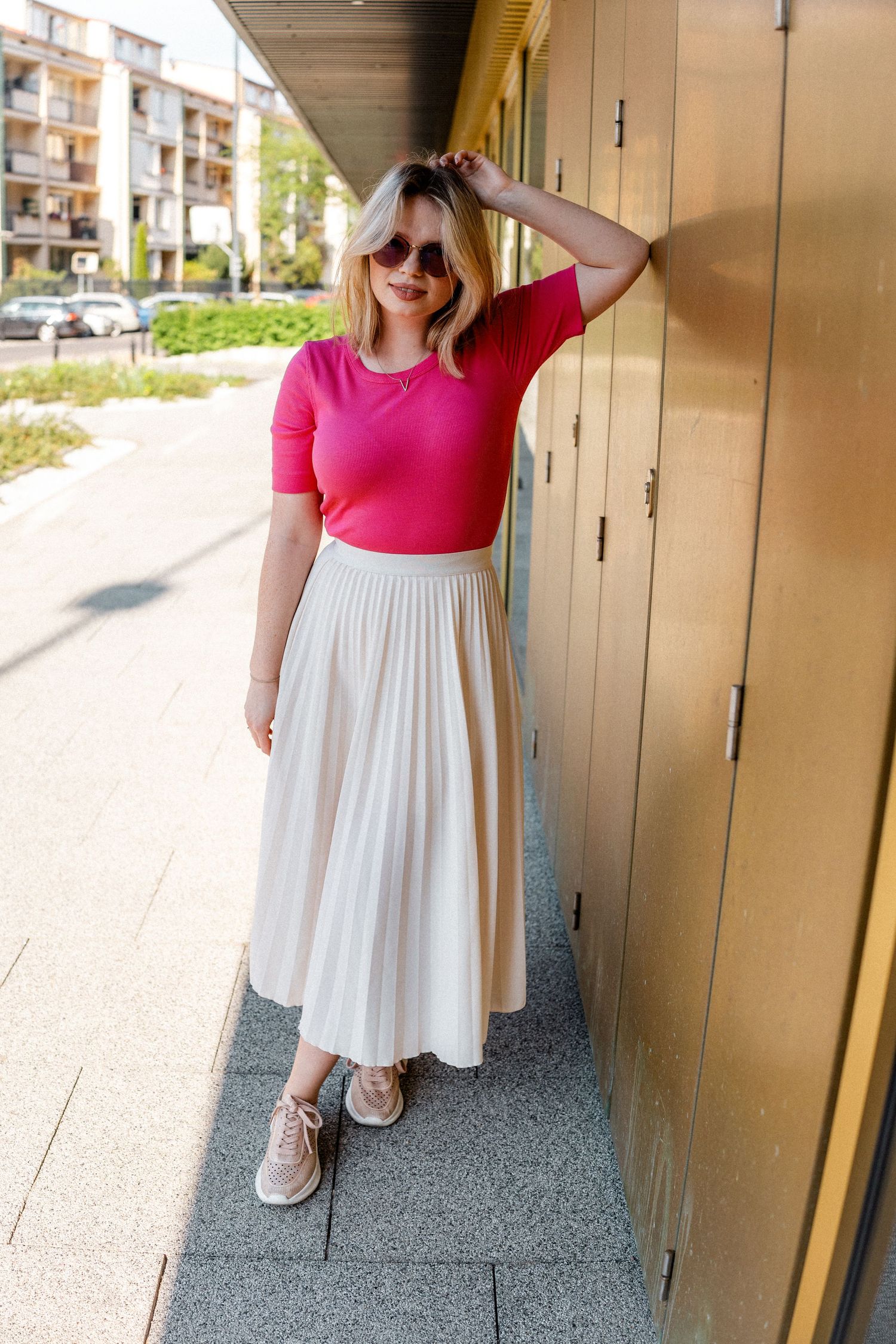 Person in pink top and white skirt posing against outdoor wall.