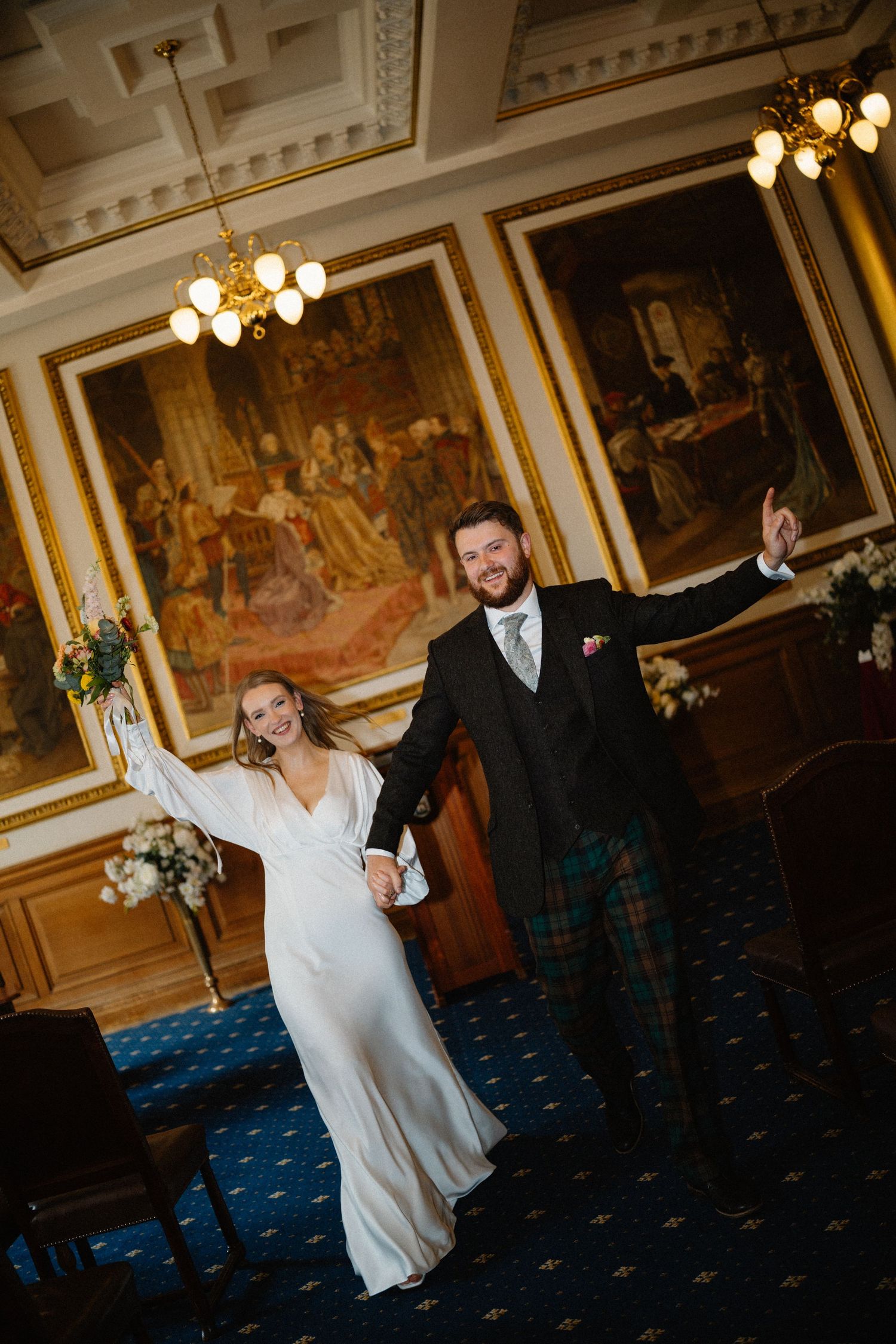 A wedding couple dances and celebrates in an ornate ballroom with vintage paintings and chandeliers.