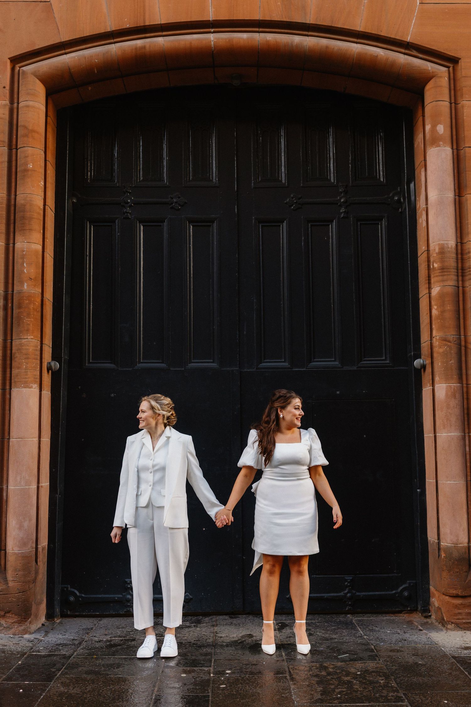 Two brides in white outfits holding hands before a large black wooden door in a stone archway.