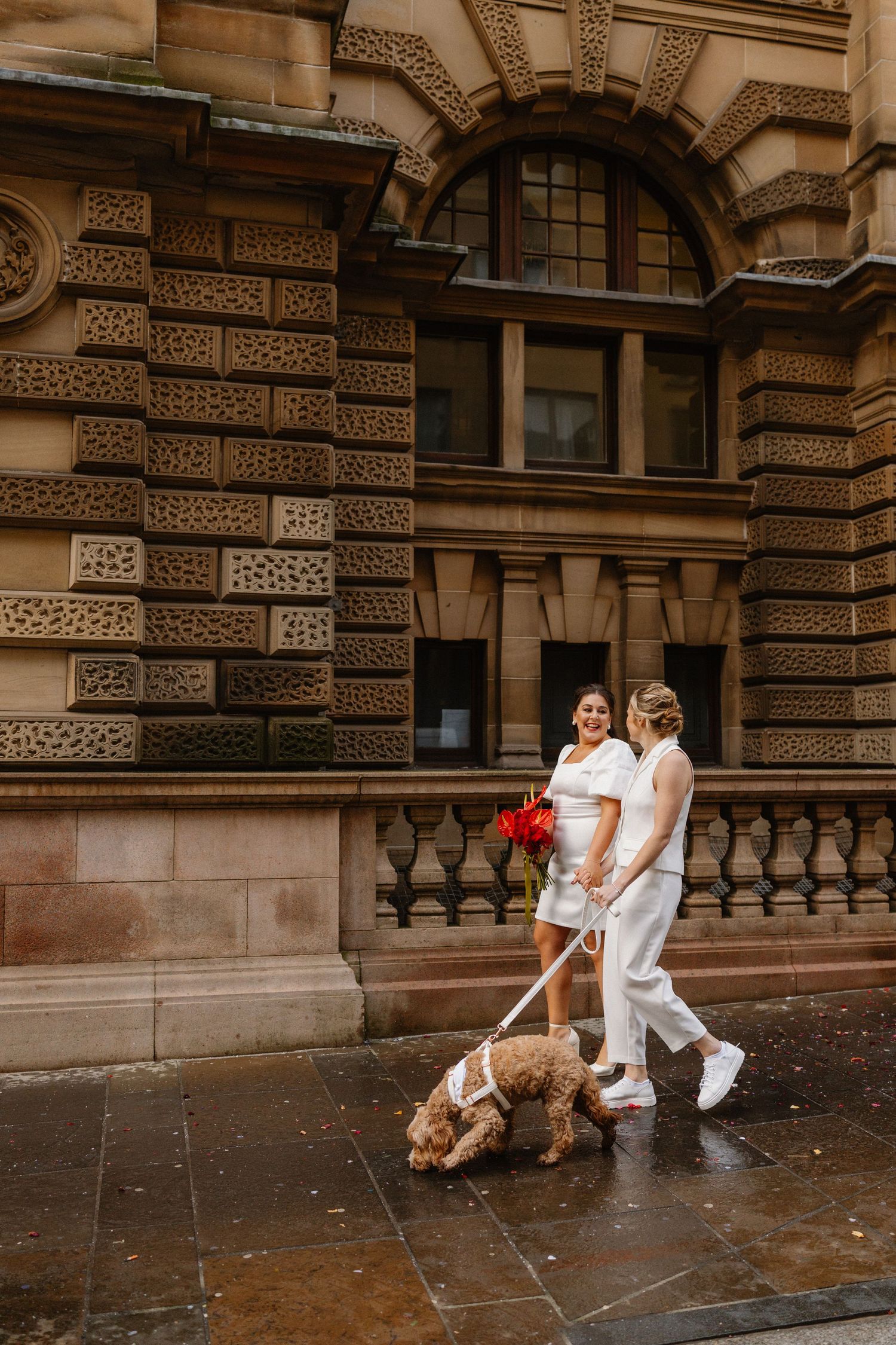 Two brides in white walk a fluffy dog past a grand sandstone building on a rainy city street.