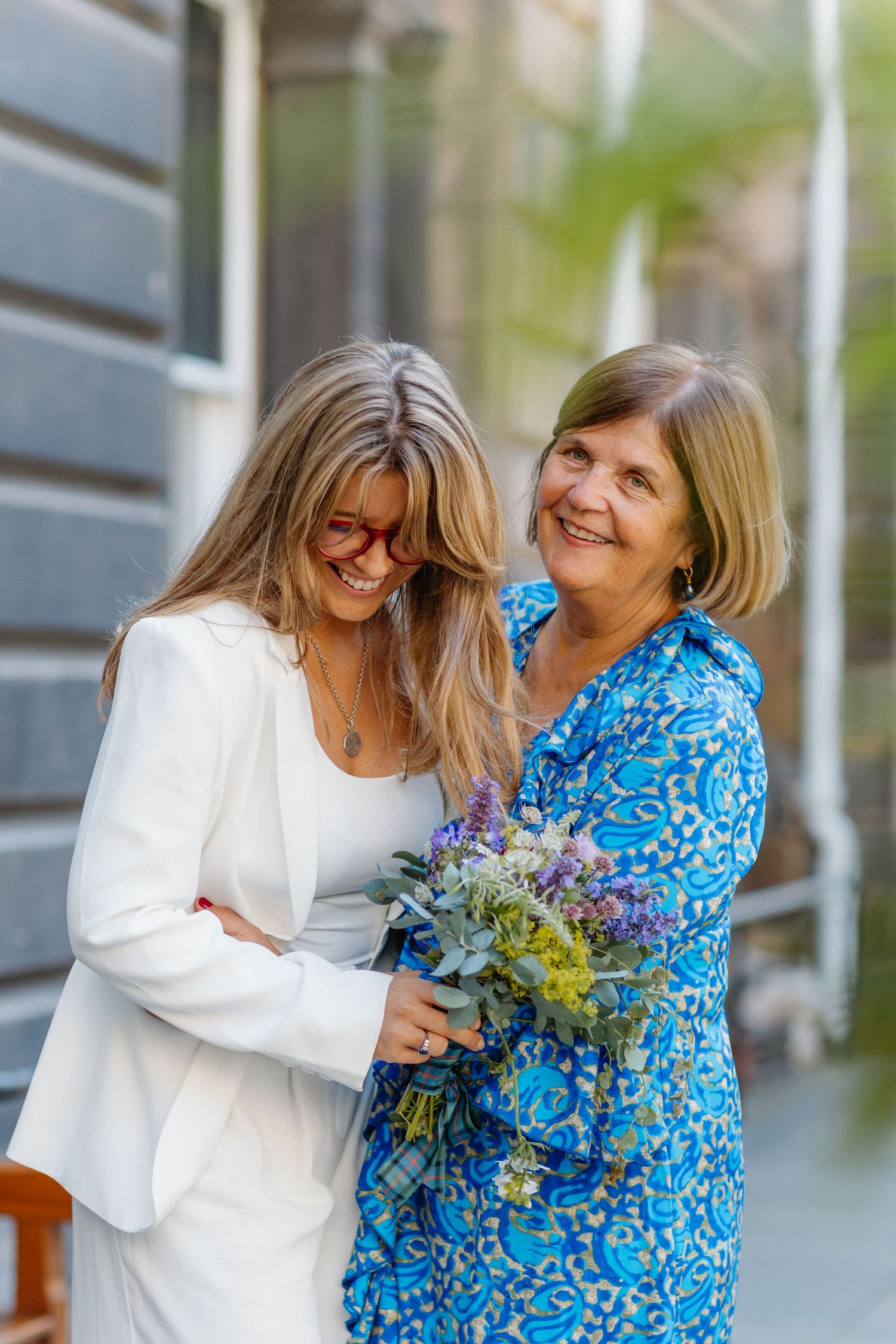 Bride in white suit laughing with mother holding wildflower bouquet outdoors.