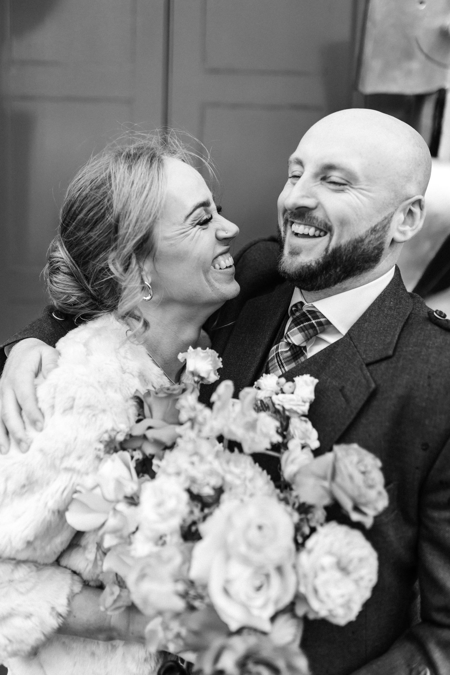 Black and white photo of laughing bride and bearded groom embracing, with bouquet of roses in foreground.