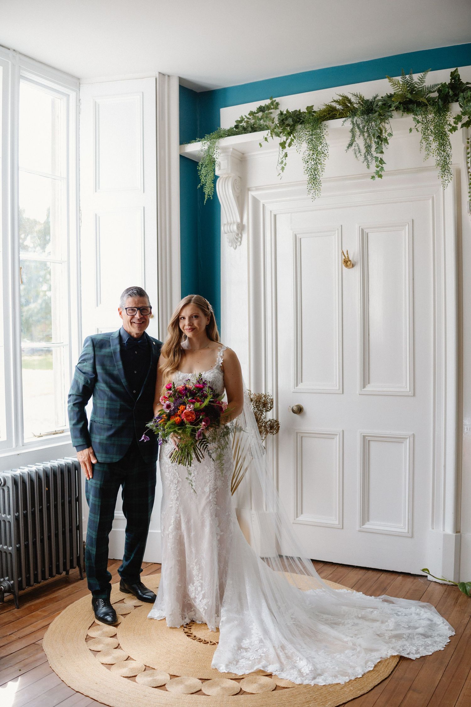 Bride in lace wedding gown holding colorful bouquet stands with suited man before white door with greenery.