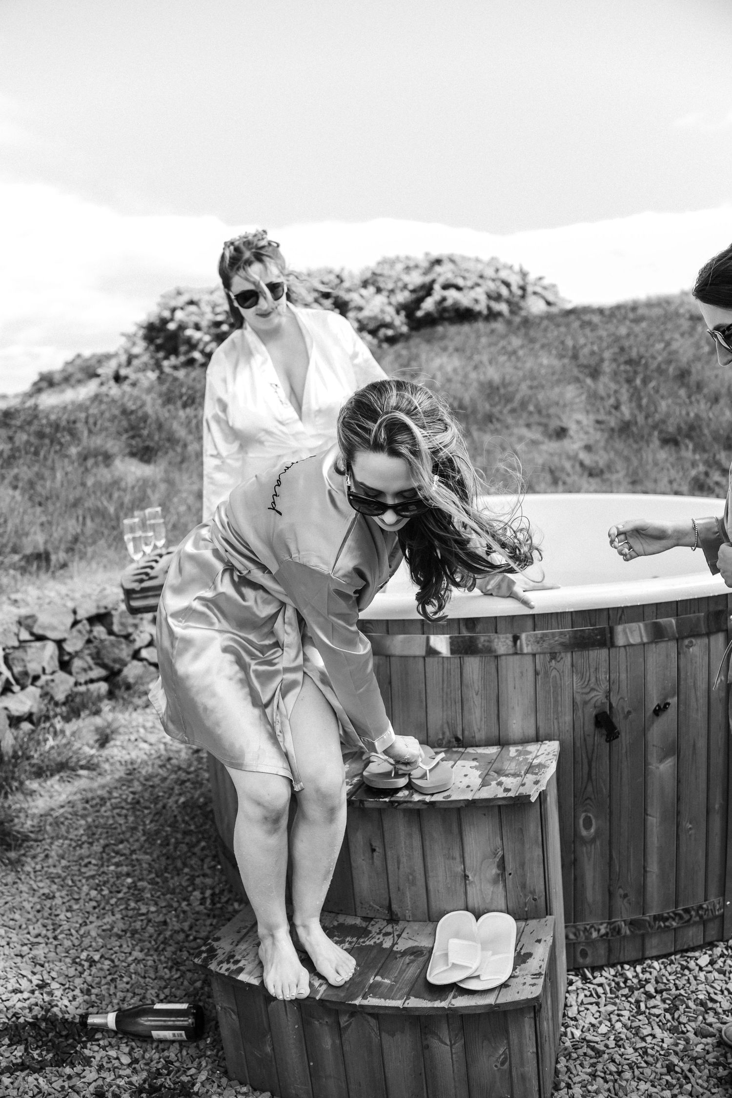 Bridesmaids in satin robes laugh near a wooden hot tub outdoors in black and white photo.