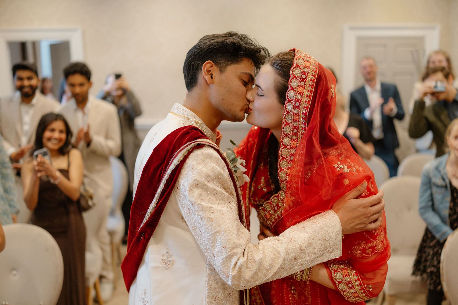 Bride in red dupatta and groom in white sherwani share first kiss at indoor wedding ceremony.