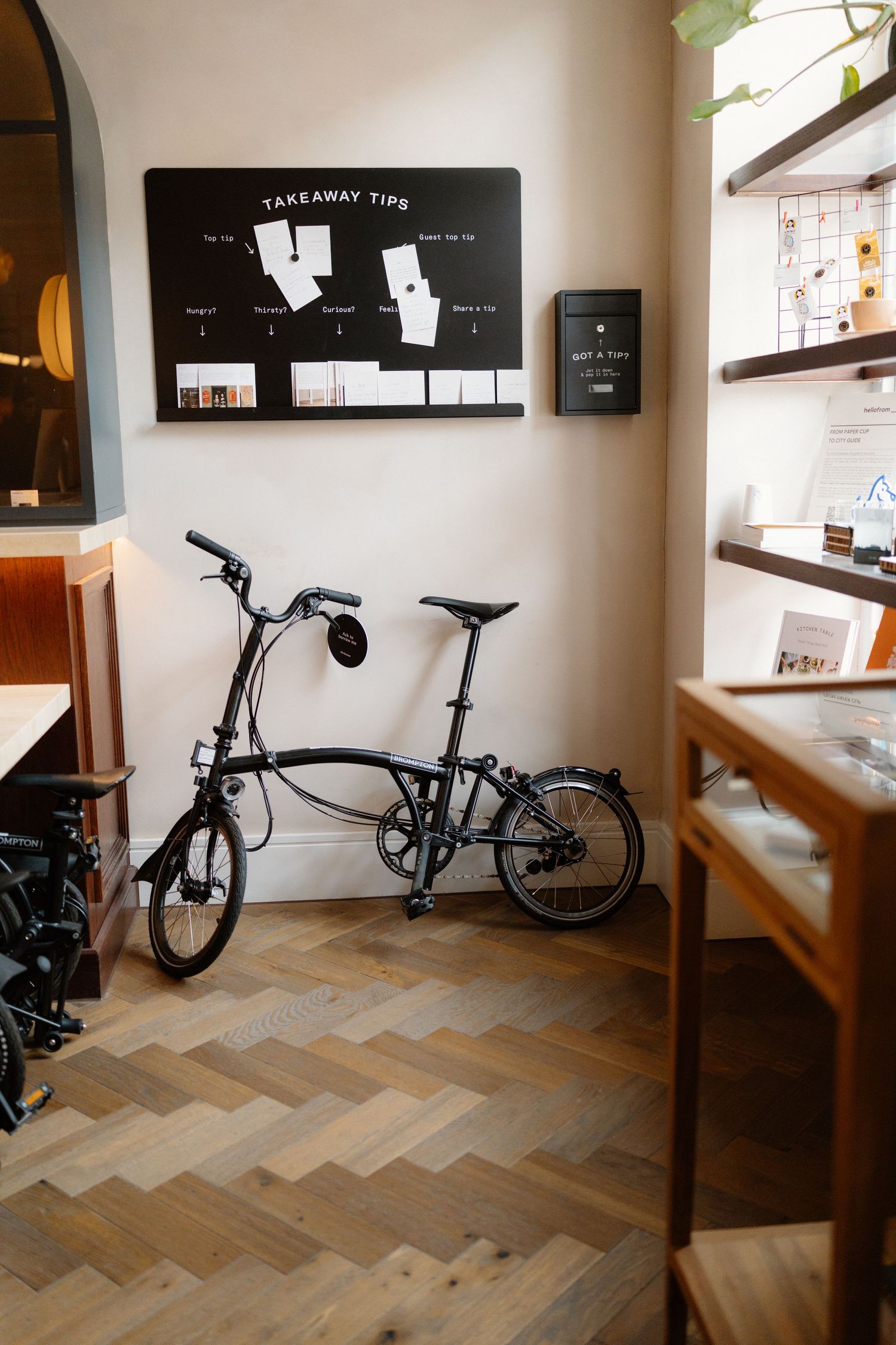 A folding Brompton bicycle parked inside a cozy cafe with herringbone wood floors and a takeaway tips chalkboard.