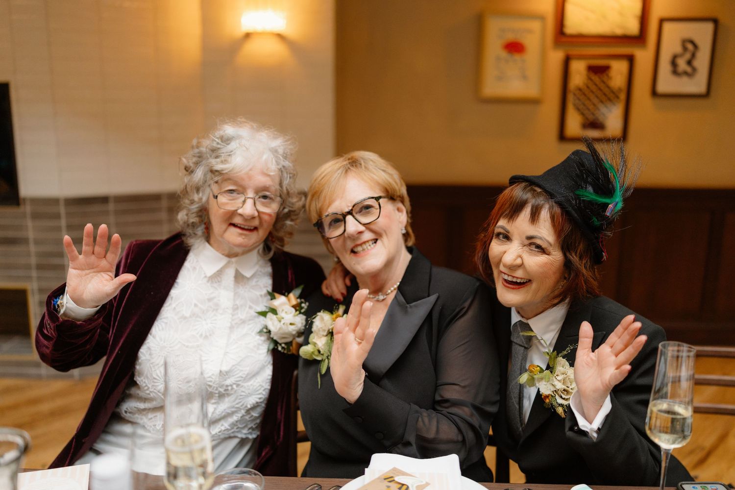 Three cheerful women waving and smiling together at a wedding reception dinner table.