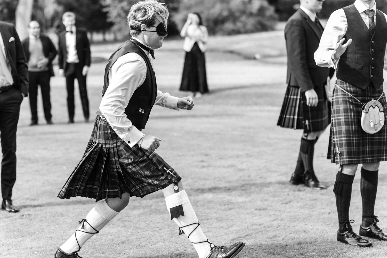 Man in traditional Scottish tartan kilt and white spats walking at outdoor wedding ceremony.