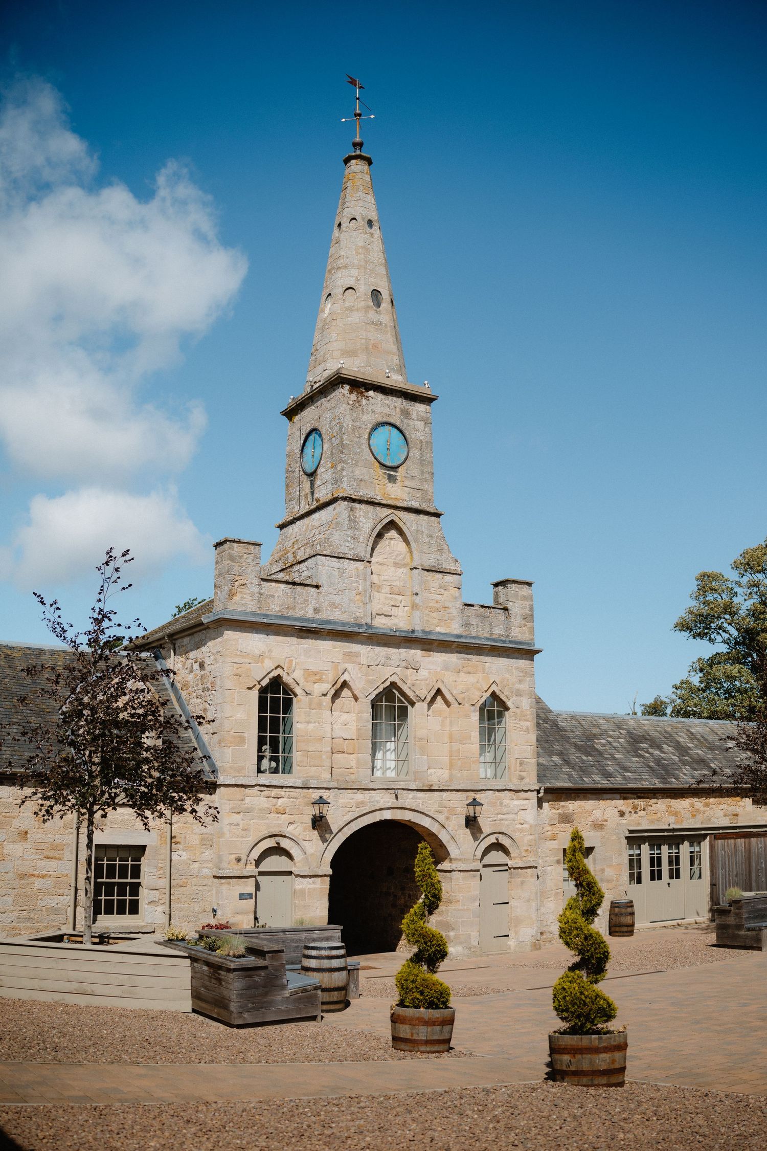 Historic stone clock tower with Gothic arched entrance, decorative spire, and barrel planters under a blue sky.