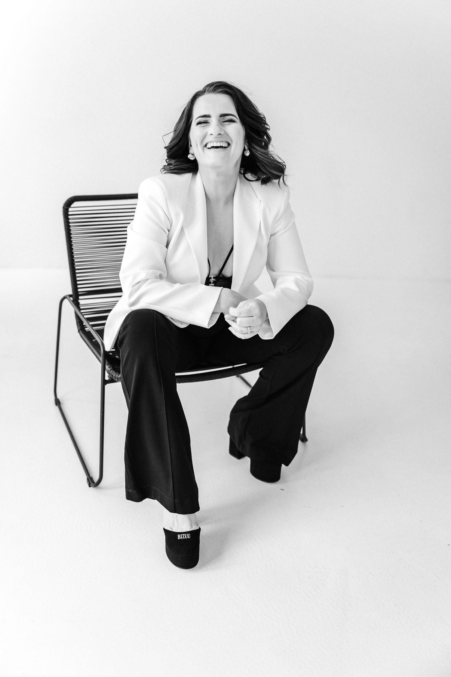 Woman in white blazer and black pants laughing while seated on a chair in a black and white studio photo.