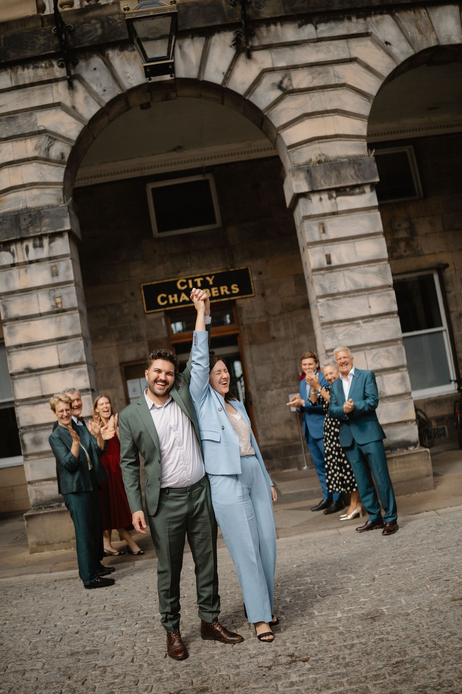 A group of people celebrating outside a historic stone building with arched architecture and a street sign.