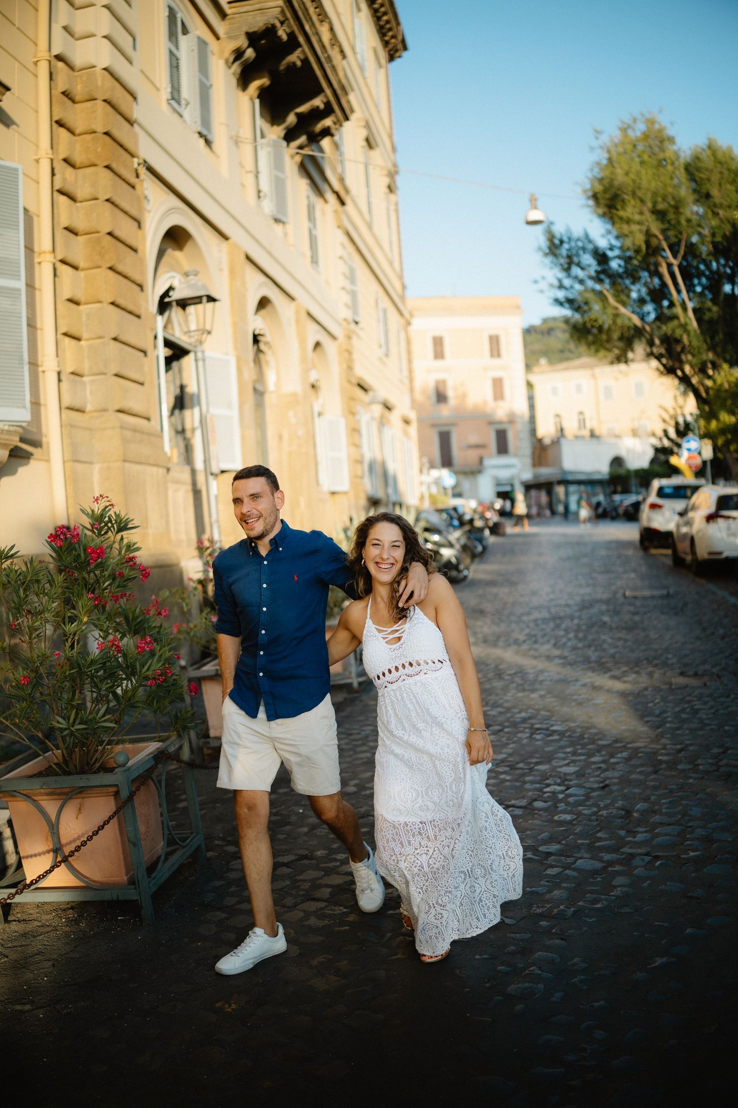 Romantic couple strolling along a sunlit cobblestone street in Italy with historic architecture.