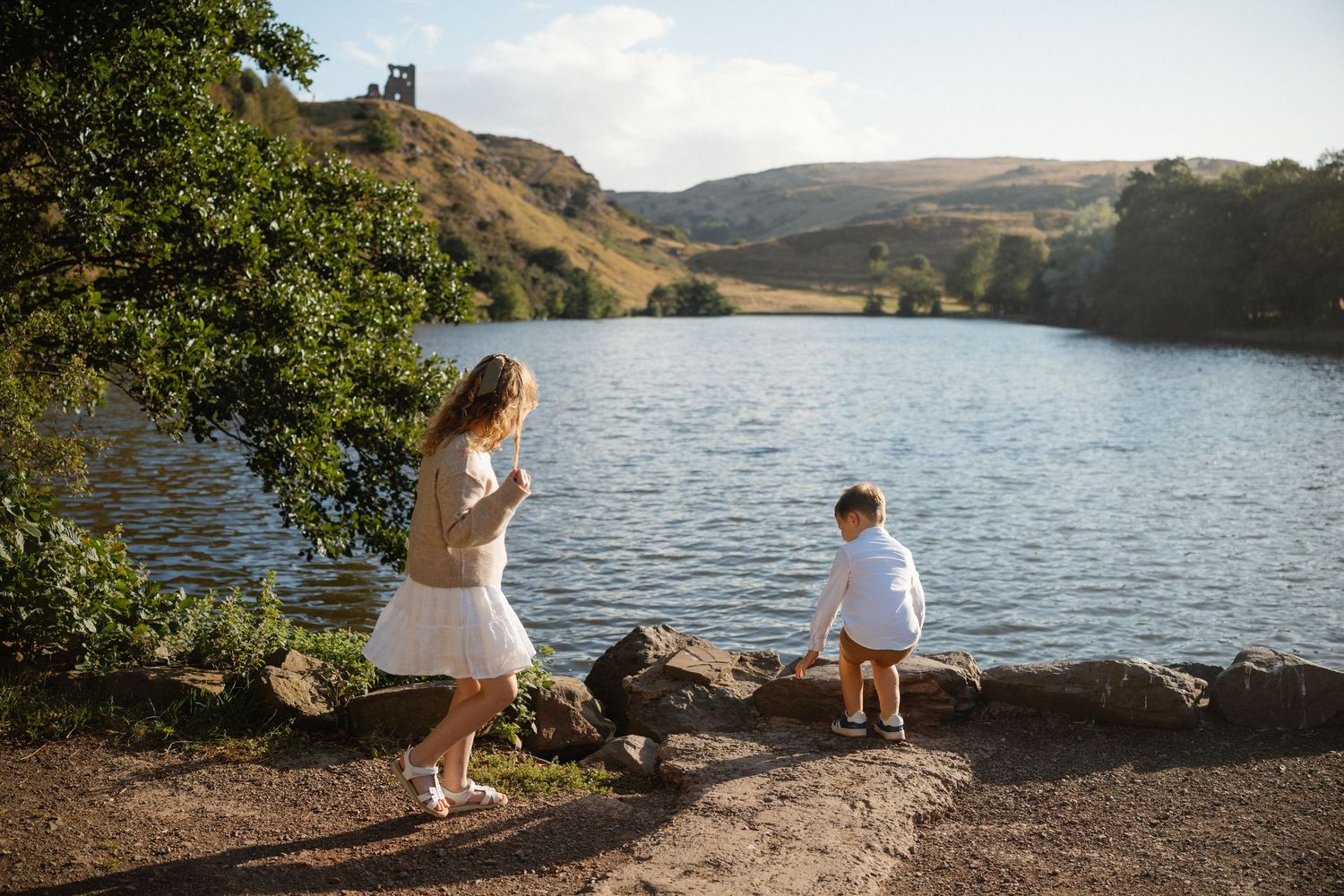 People exploring rocky lakeside with mountains in the background on a sunny day.