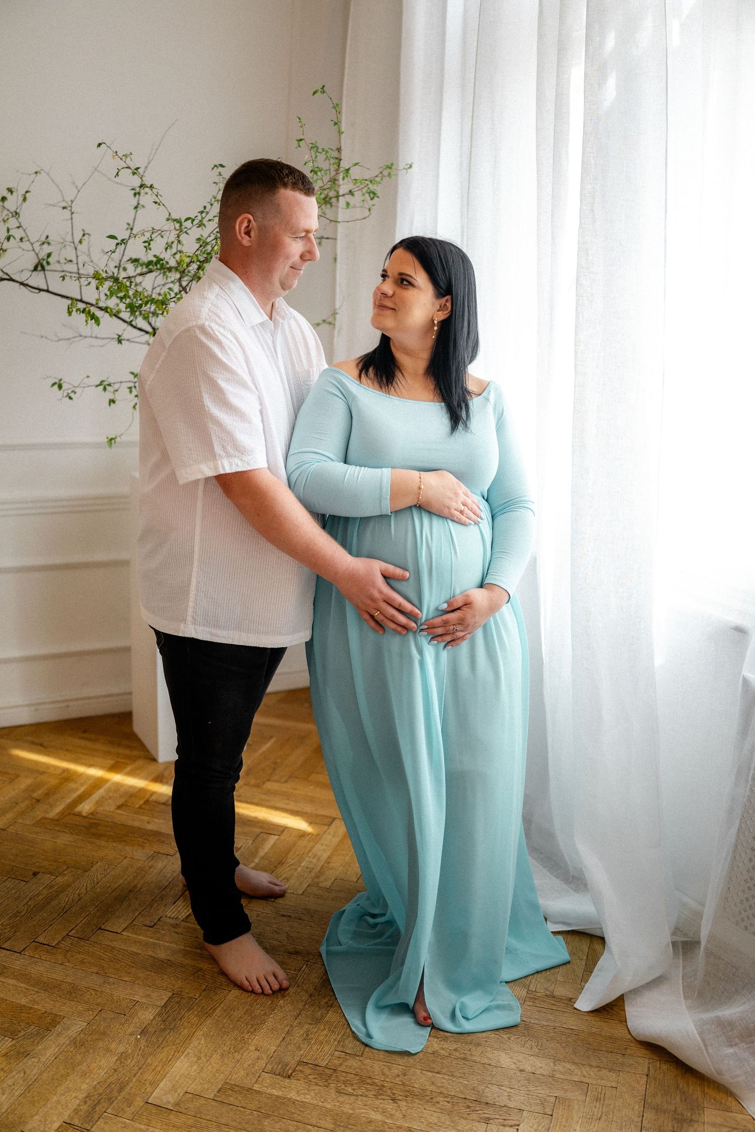 Couple in light blue and white outfits posing together in bright indoor setting with hardwood floors.