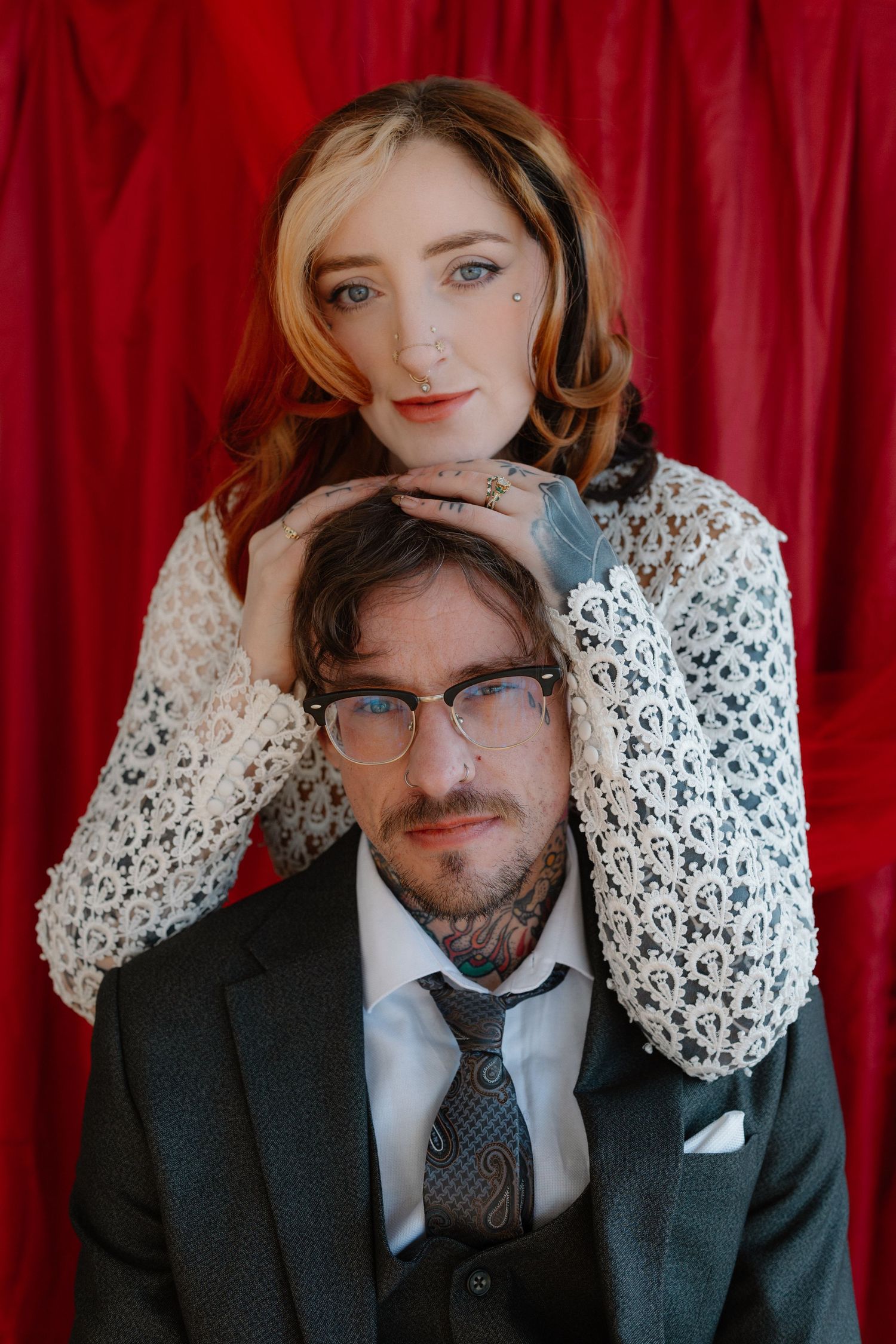 Couple posing against red curtain, woman in lace top resting hands on mans head, man in suit with neck tattoos and glasses.