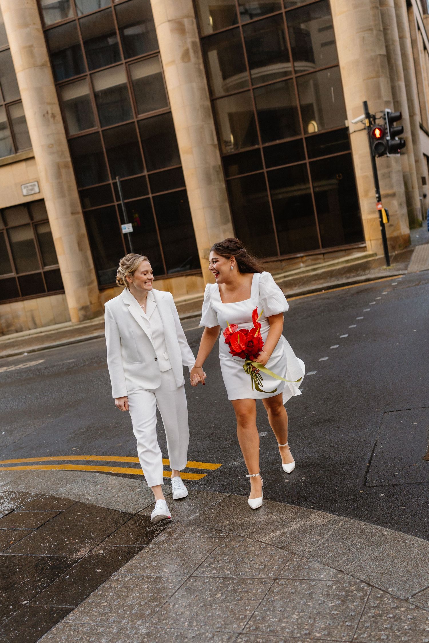Two brides in white outfits hold hands crossing a city street, one carrying a red bouquet.