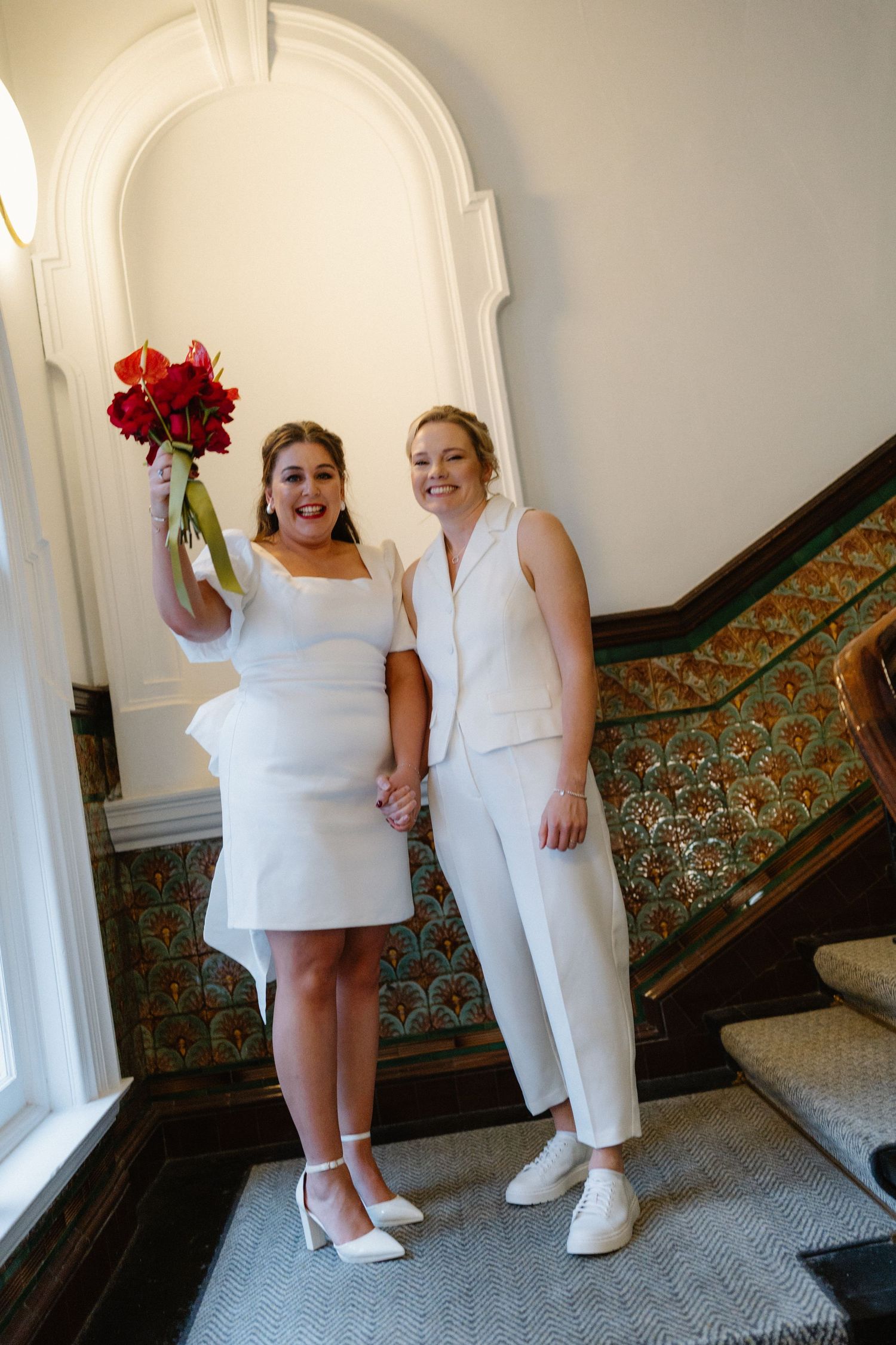 Two brides in white attire hold hands and celebrate on a staircase, one raising a red bouquet.