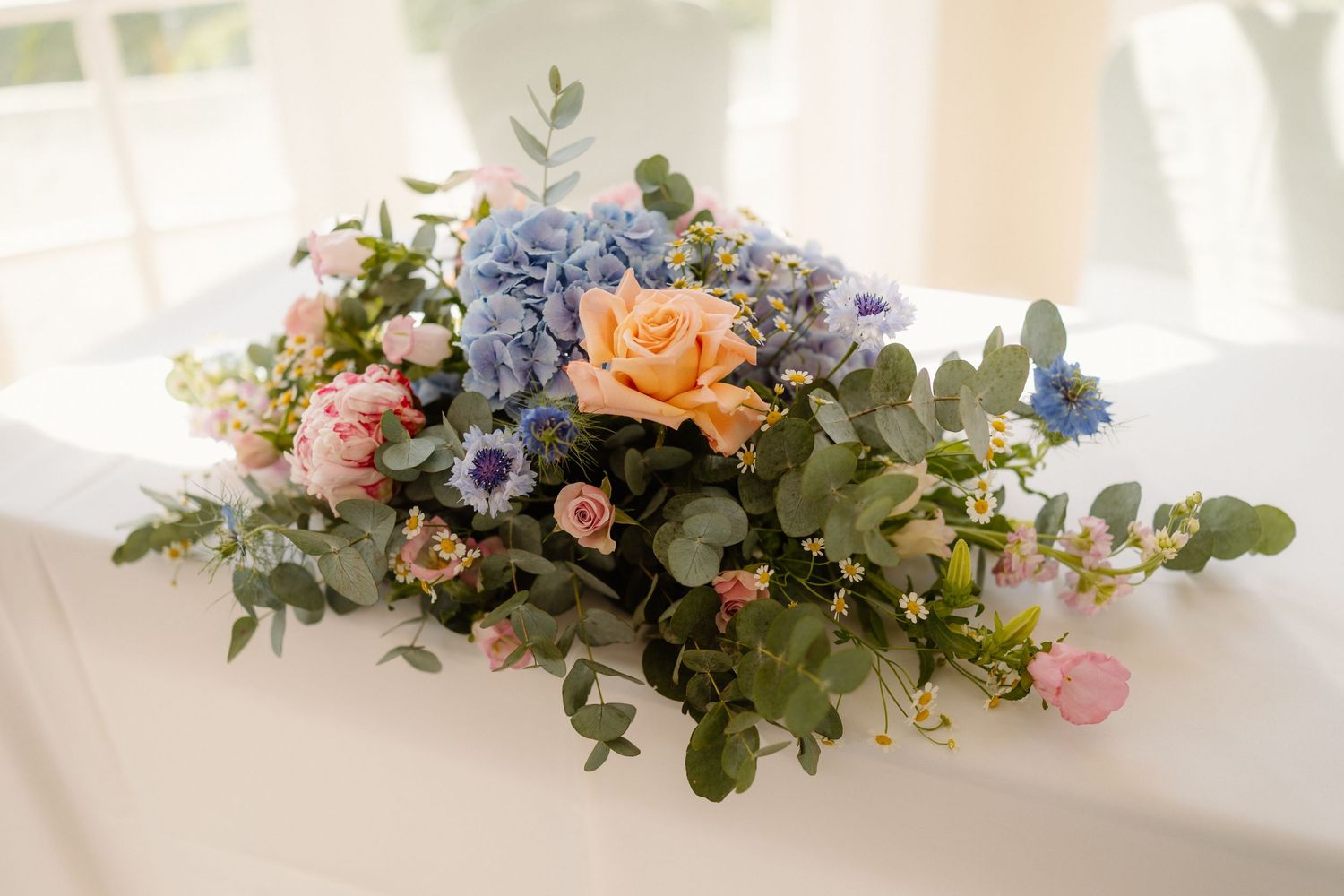 A elegant floral arrangement of blue hydrangeas, peach roses, and pink blooms with eucalyptus on a white surface.