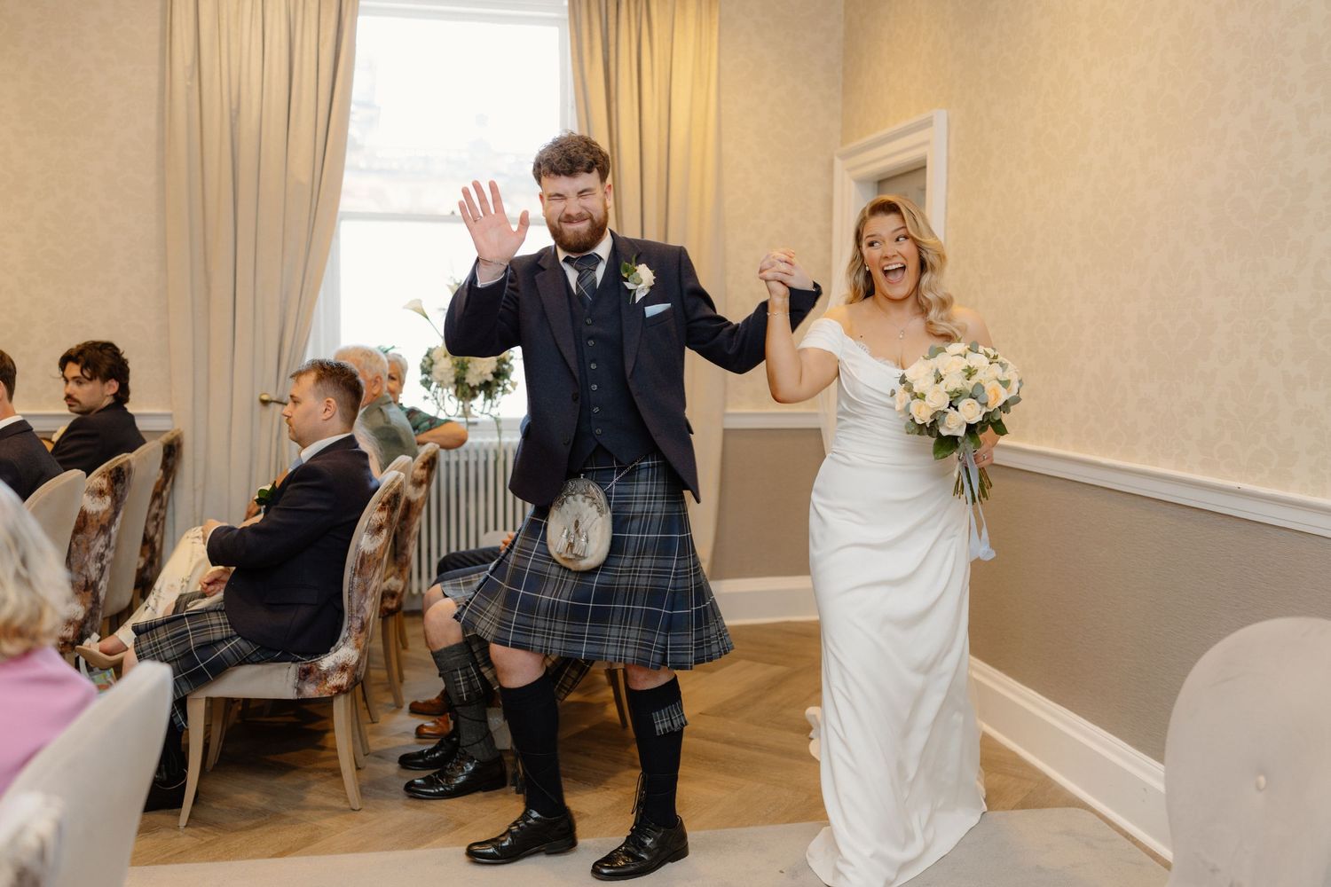 Joyful newlyweds walk down the aisle, groom in Scottish kilt and bride in white gown holding bouquet at wedding ceremony.