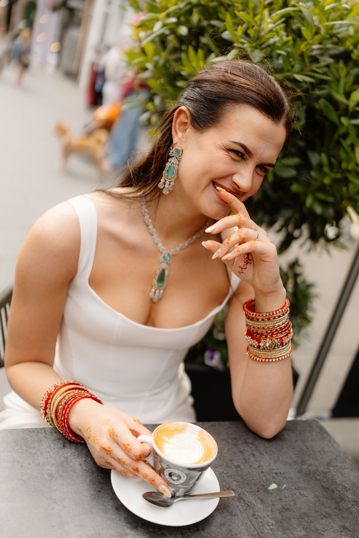 A smiling woman in a white dress with henna hands and colorful jewelry enjoys a latte at an outdoor cafe.