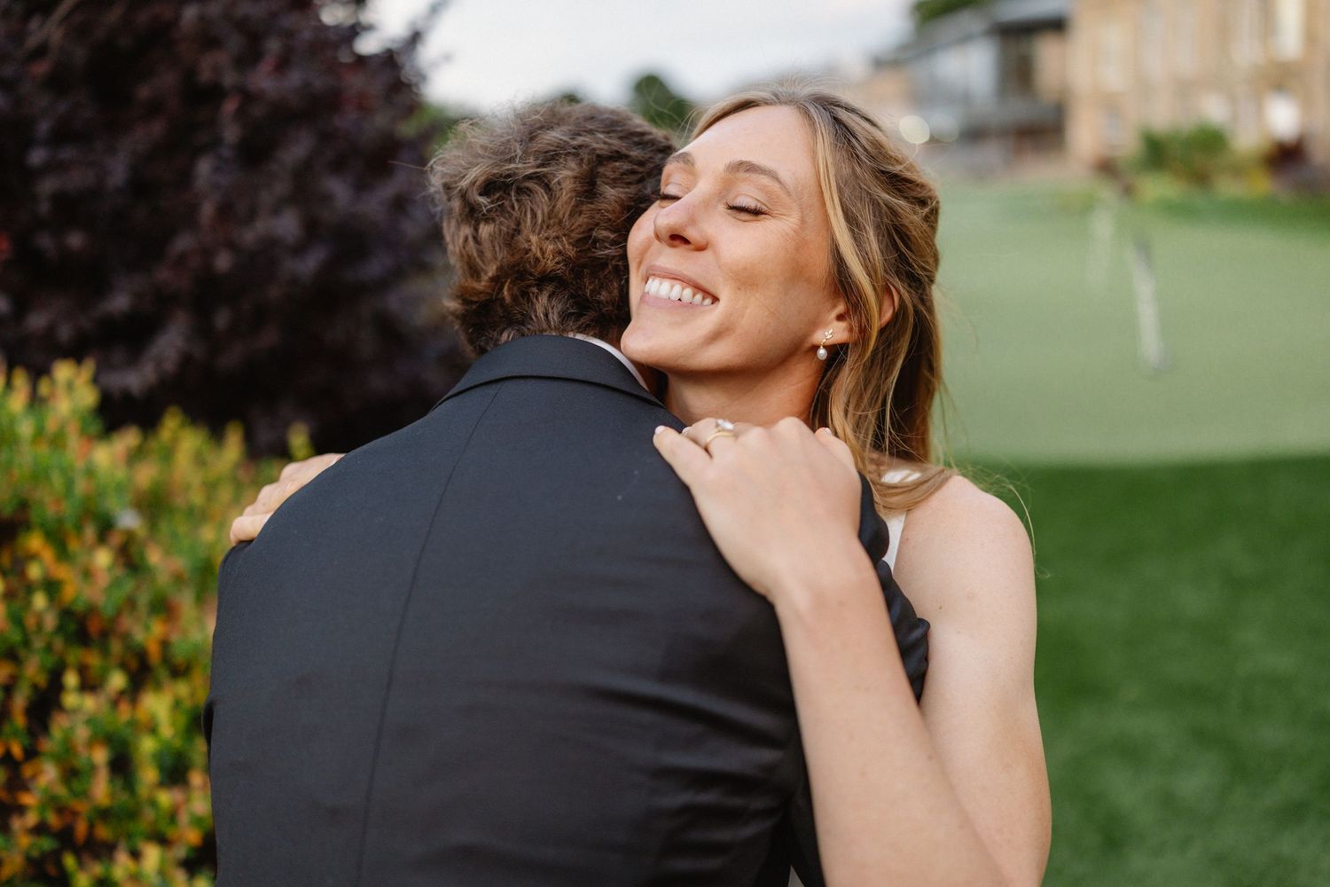A smiling bride hugs her groom outdoors on a green lawn during a romantic engagement or wedding celebration.