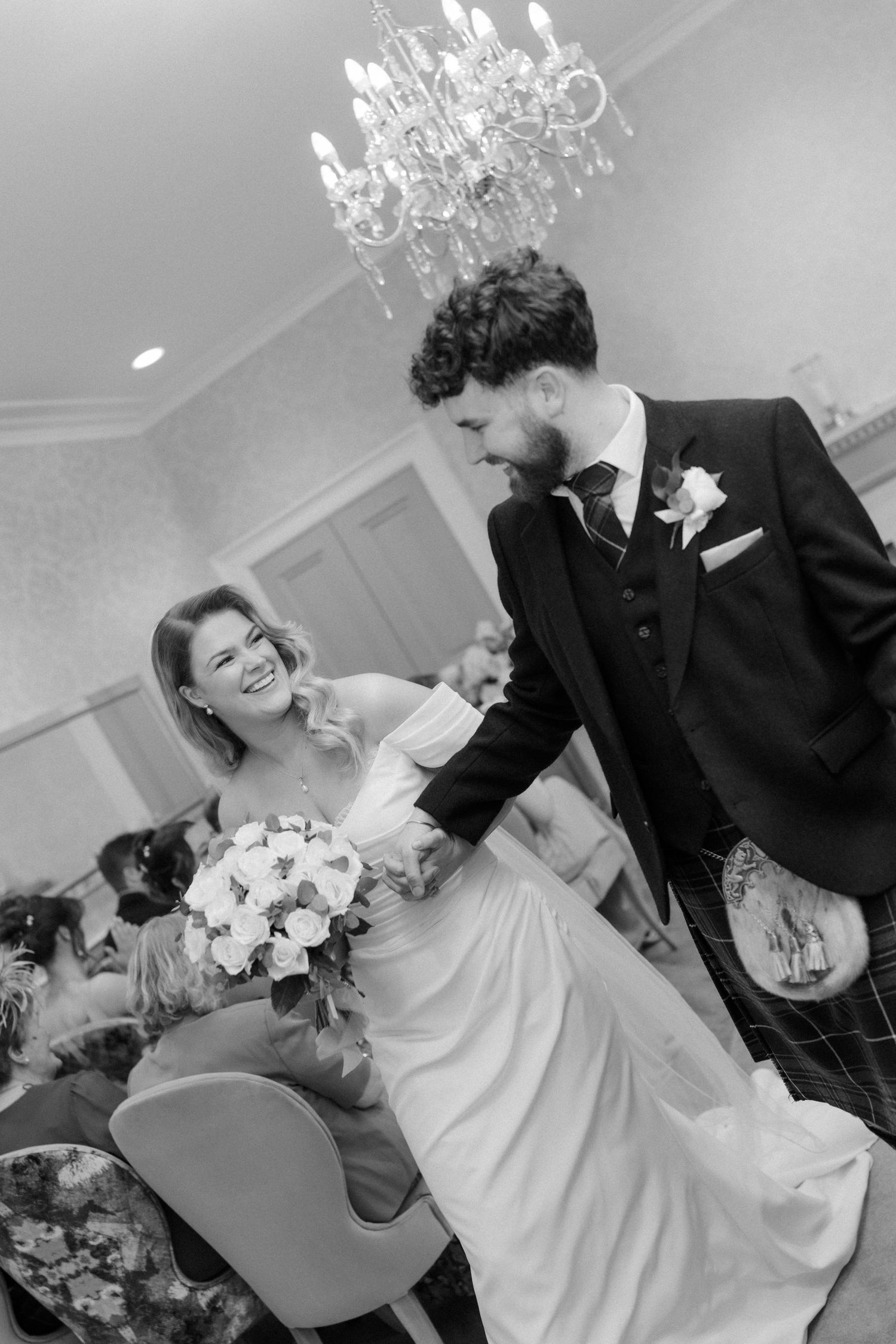 Bride and groom share joyful moment at wedding reception under elegant crystal chandelier in black and white.