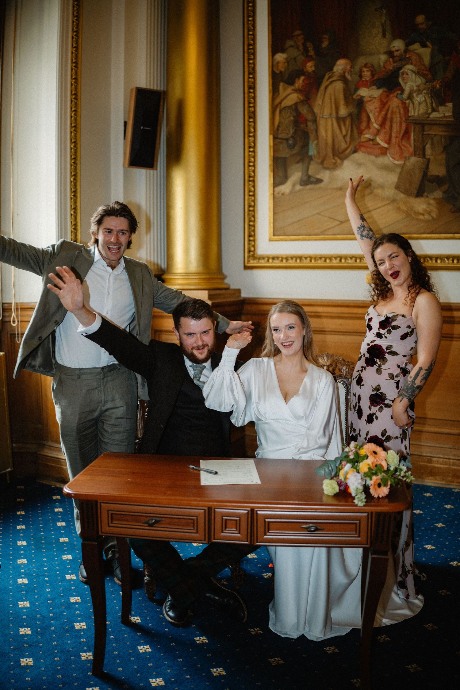 Joyful newlyweds signing wedding register at ornate desk with celebrating witnesses in elegant room.