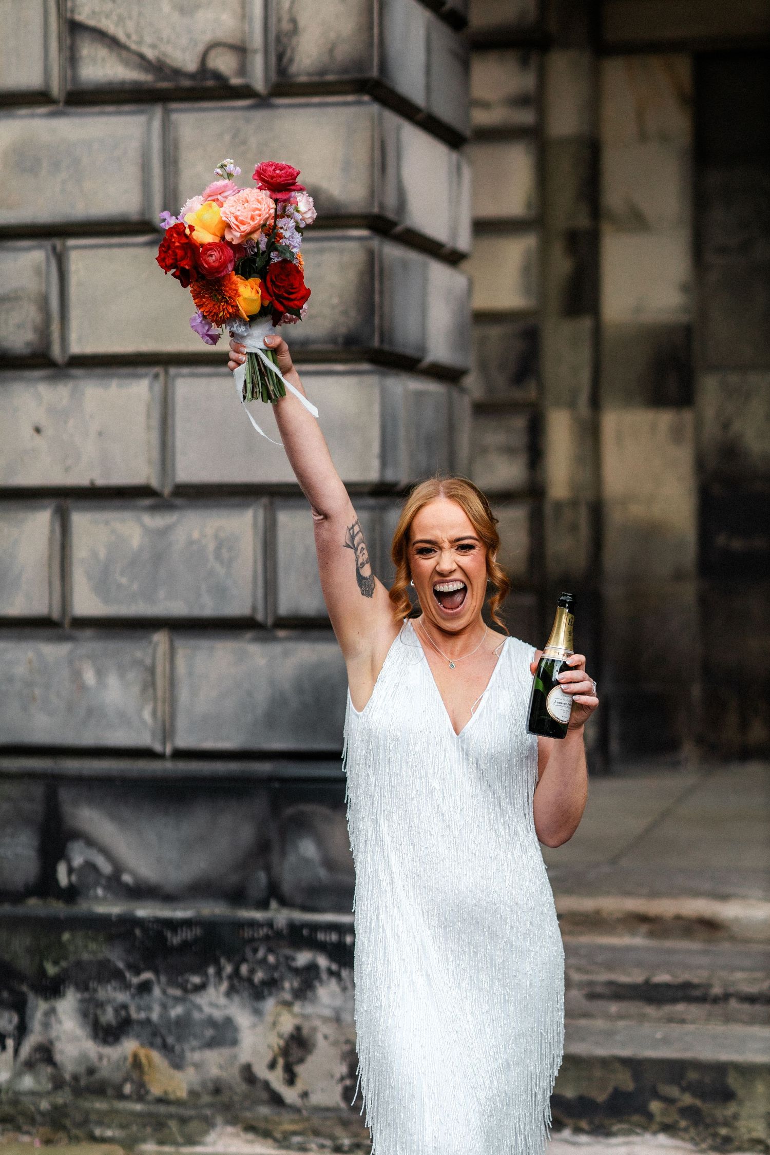 Excited bride in white fringe dress celebrating with bouquet and champagne bottle outdoors.