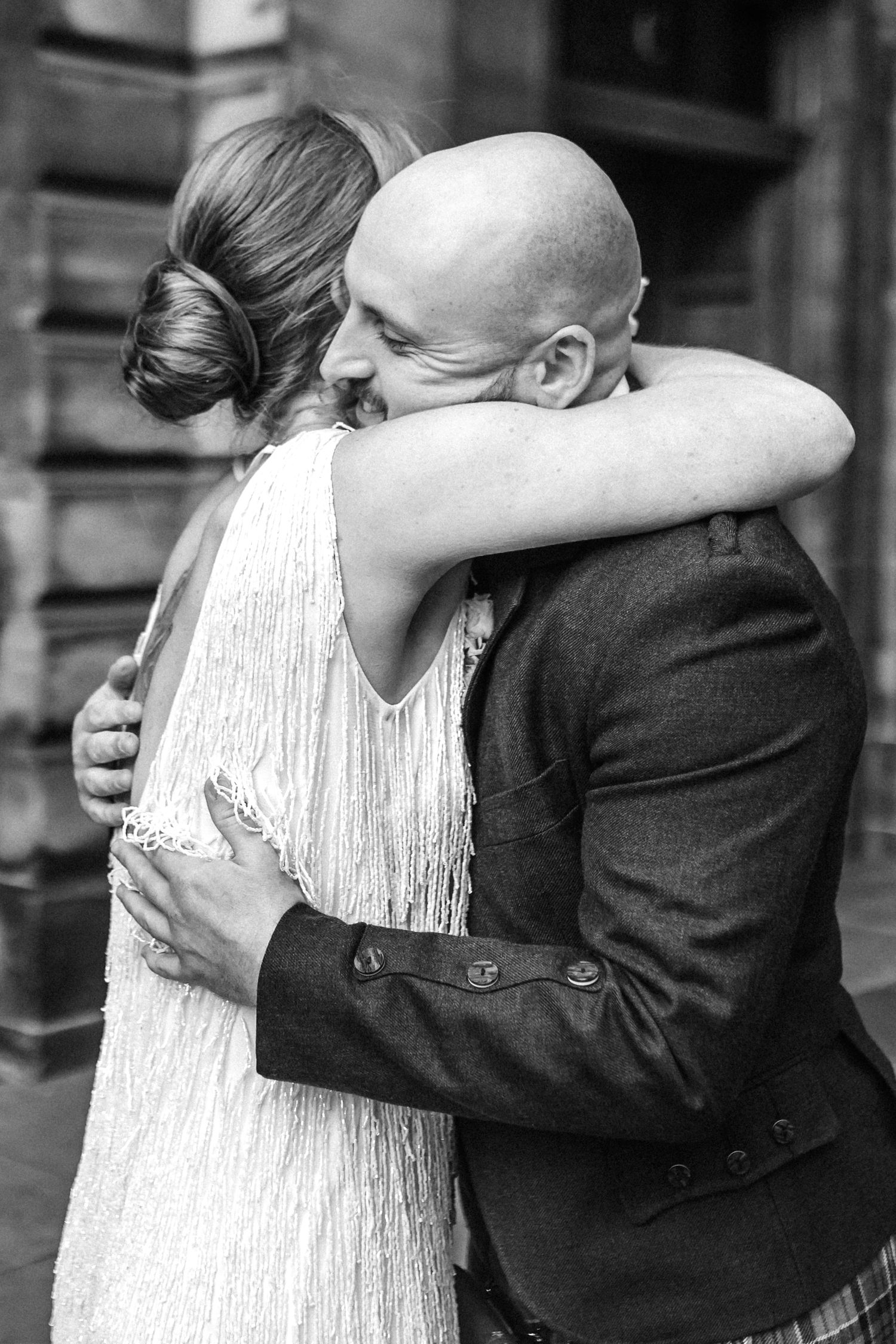 Black and white photo of bald groom in tweed suit embracing bride in fringed white dress tenderly.