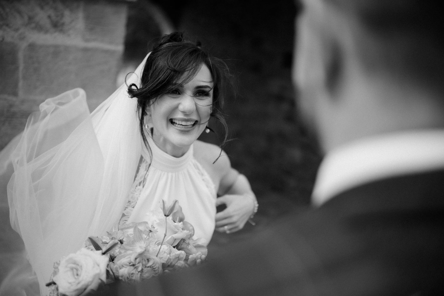 Black and white close-up of a smiling bride in a veil holding a bouquet, looking lovingly at the groom.