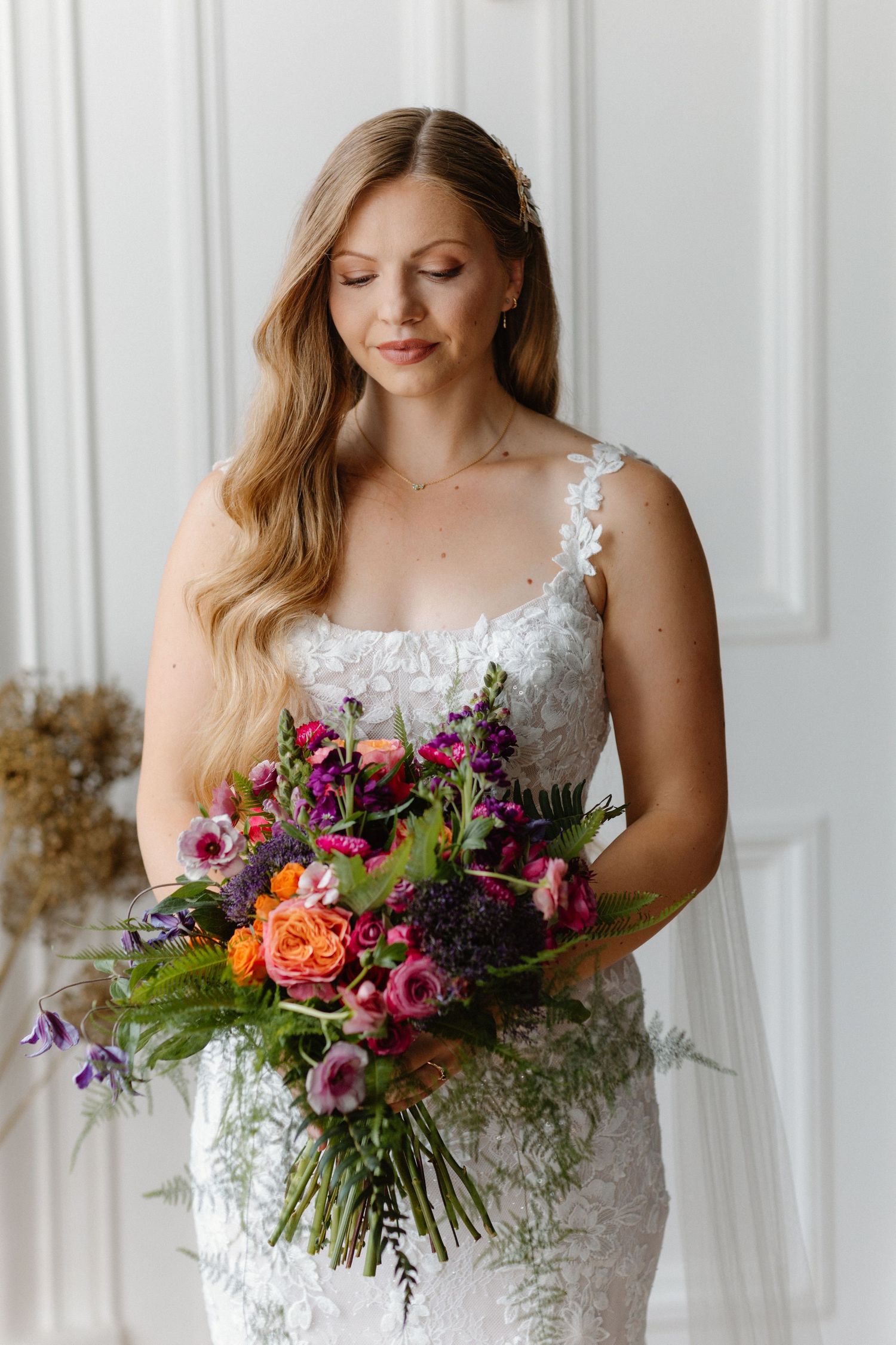 Blonde bride in lace gown holding vibrant purple, pink and orange wedding bouquet indoors.