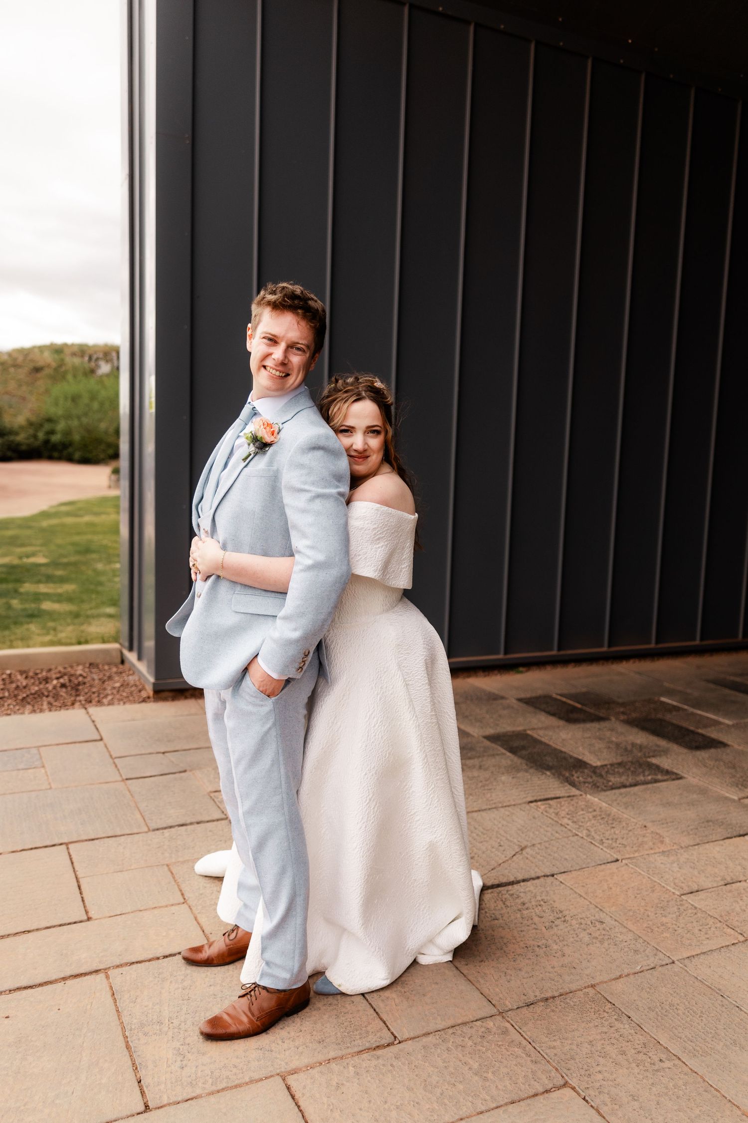 Bride in white off-shoulder gown embraces groom in light blue suit outside modern dark barn wedding venue.