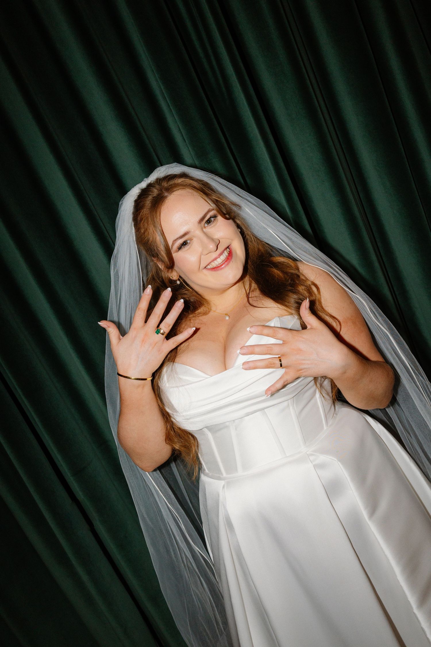 Smiling bride in white wedding dress and veil posing against green velvet curtain backdrop.