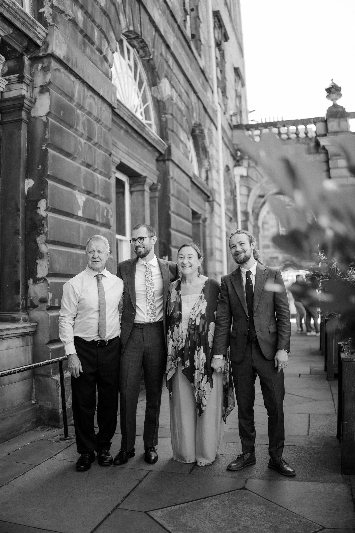 Black and white photo of four elegantly dressed people posing together outside a historic stone building.