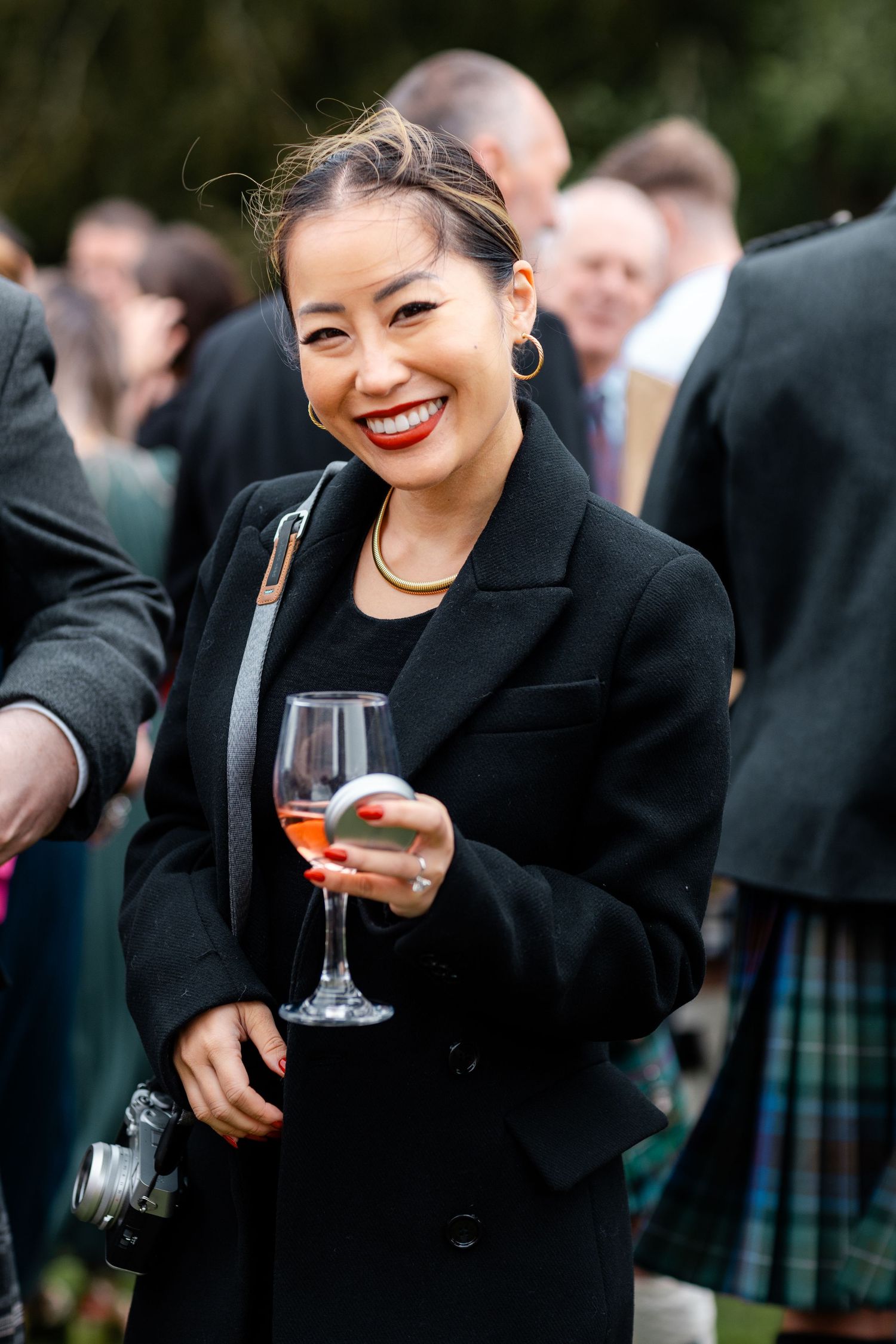 Smiling Asian woman in black coat holding a glass of rosé wine at an outdoor wedding reception.