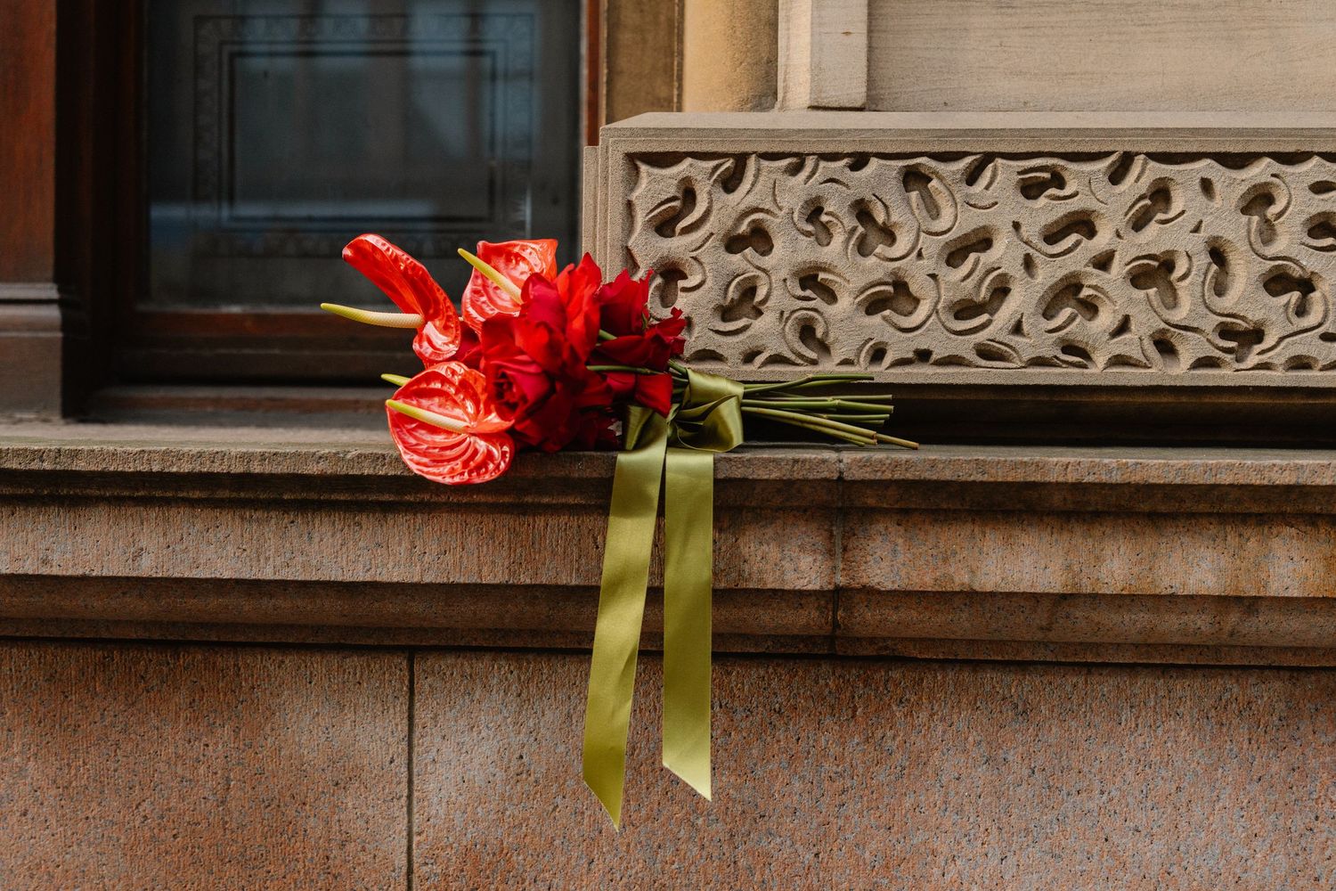 Red anthurium bouquet with olive green ribbon resting on a stone window ledge of a classic building.