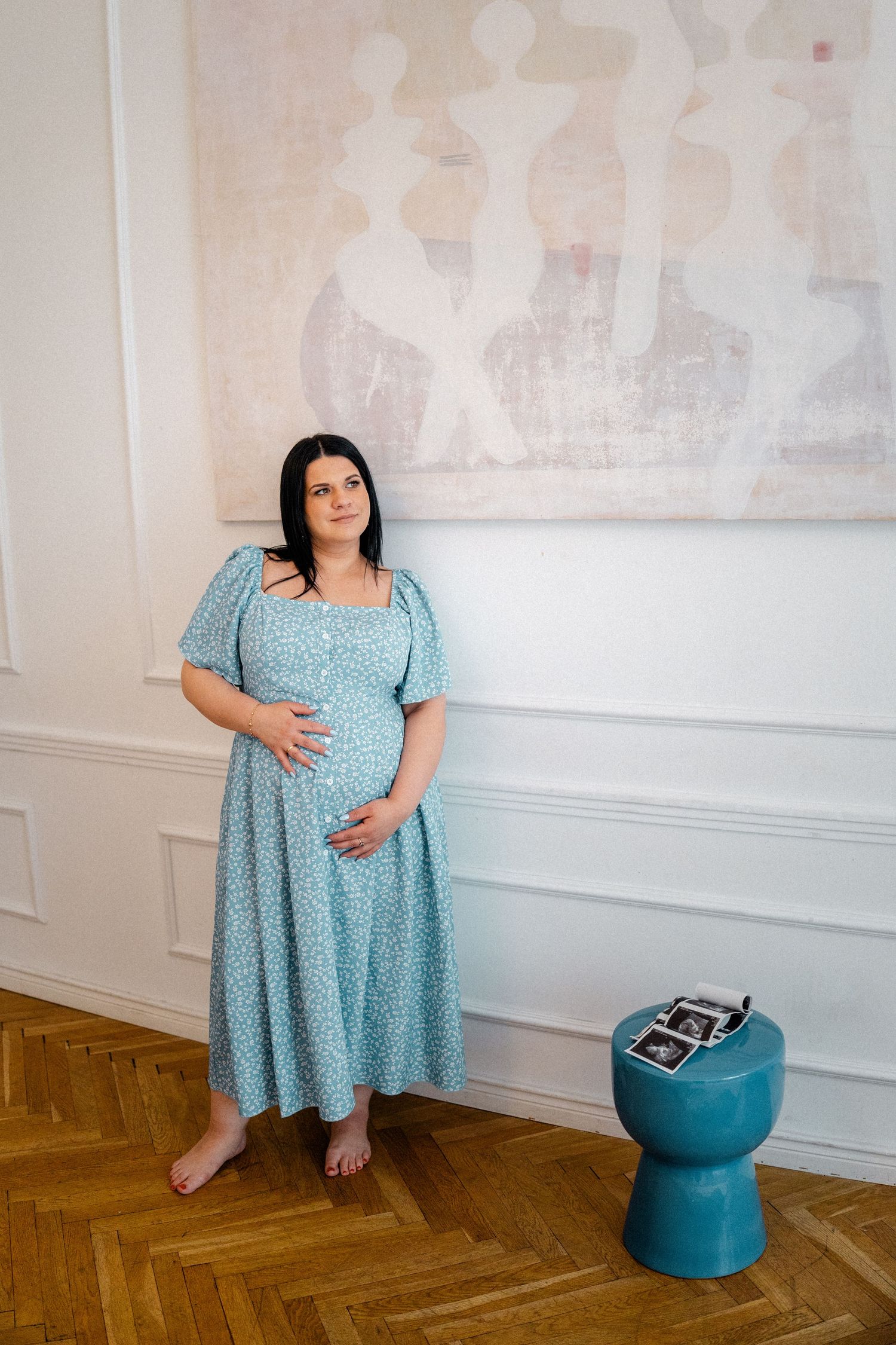 Woman in blue polka dot dress posing against white wall with decorative turquoise ottoman.