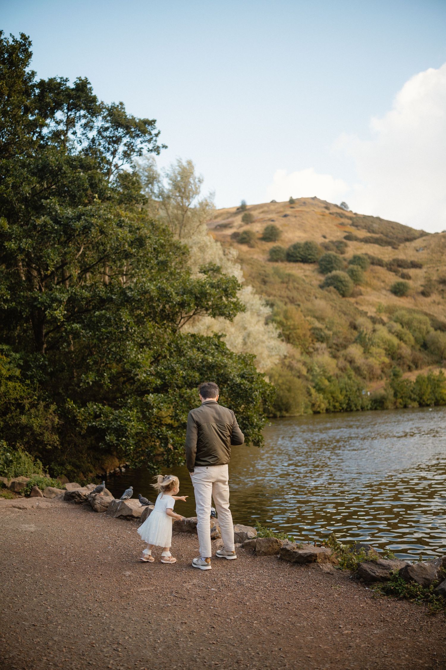 Adult and child standing together by a lake with hills in the background.
