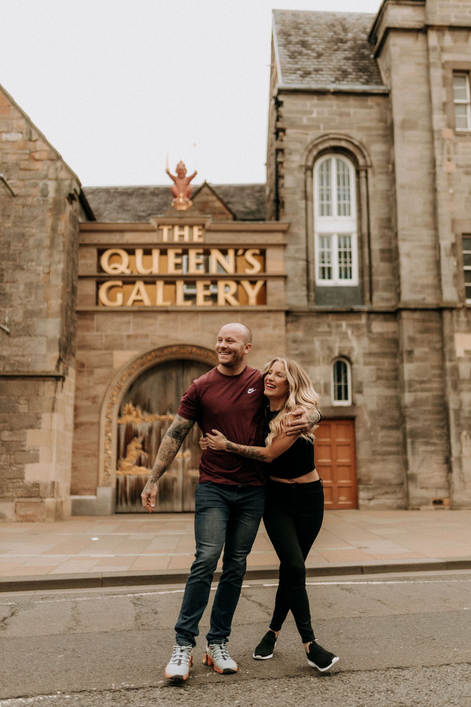 A playful couple poses and laughs together outside The Queens Gallery building with stone architecture.