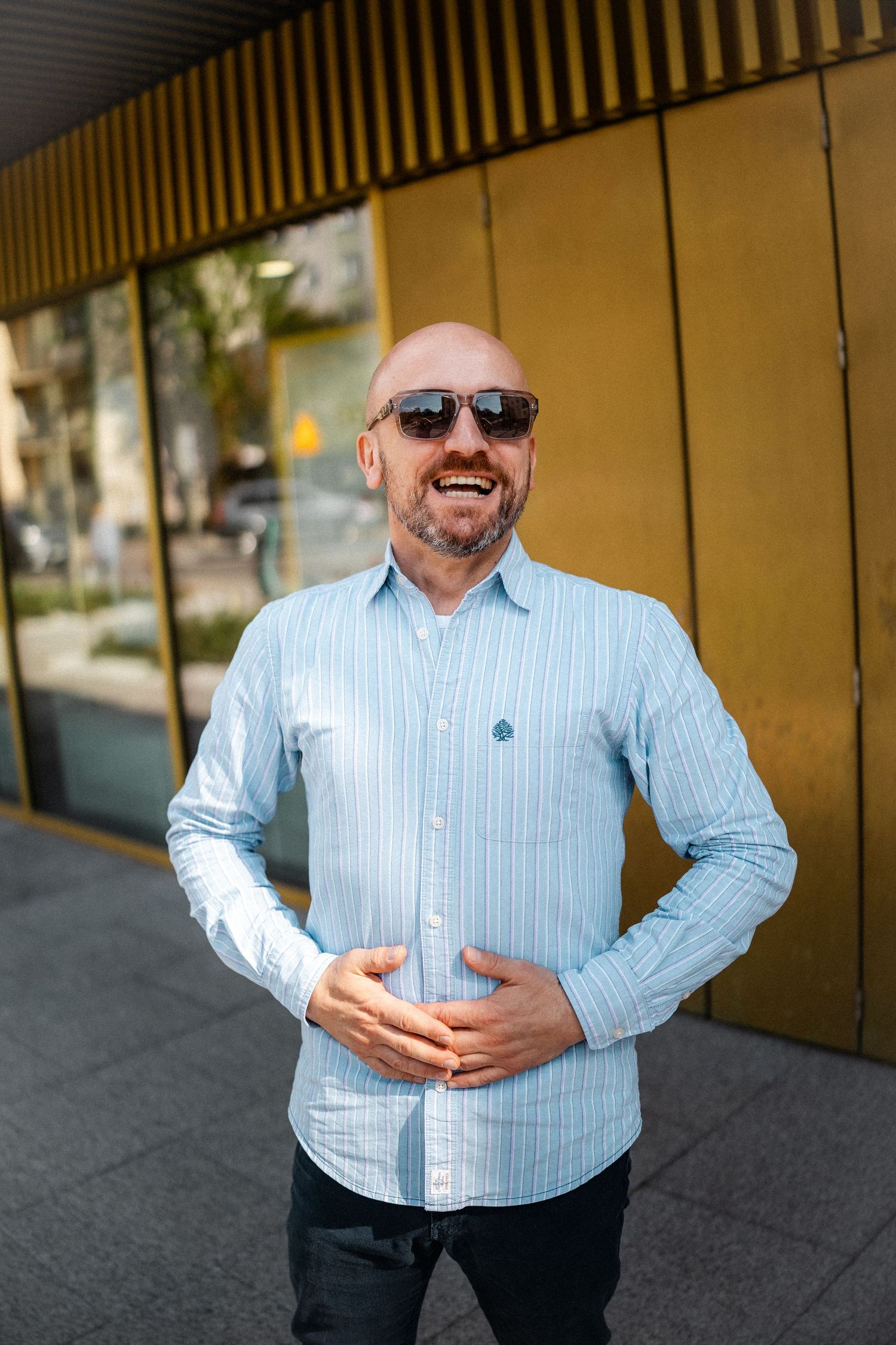 Person in light blue striped shirt and sunglasses posing against gold wall.