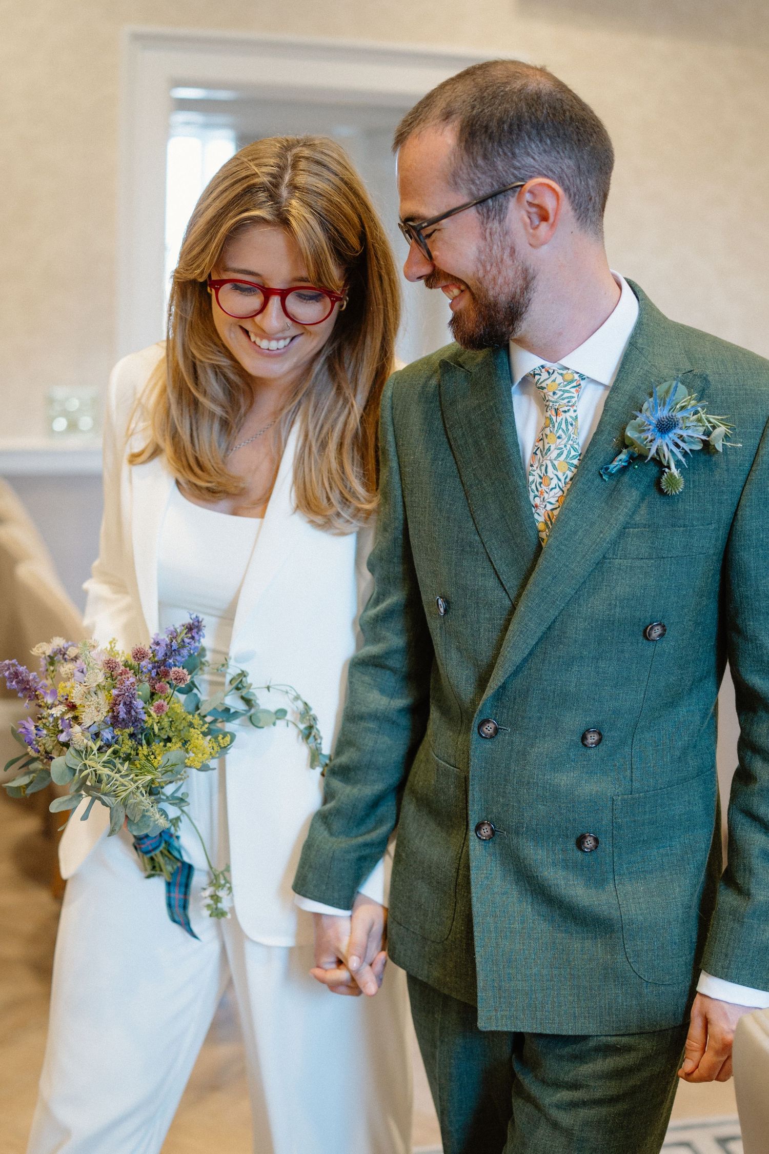 Stylish couple in white dress and green suit holding bouquet of blue and purple flowers.