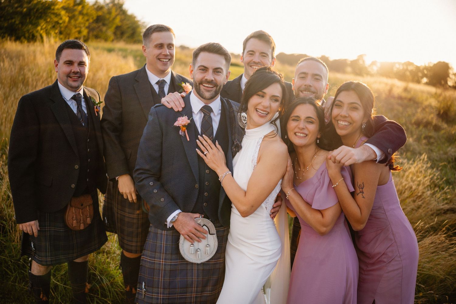 Wedding party laughs and poses together outdoors during golden hour sunset photography session.