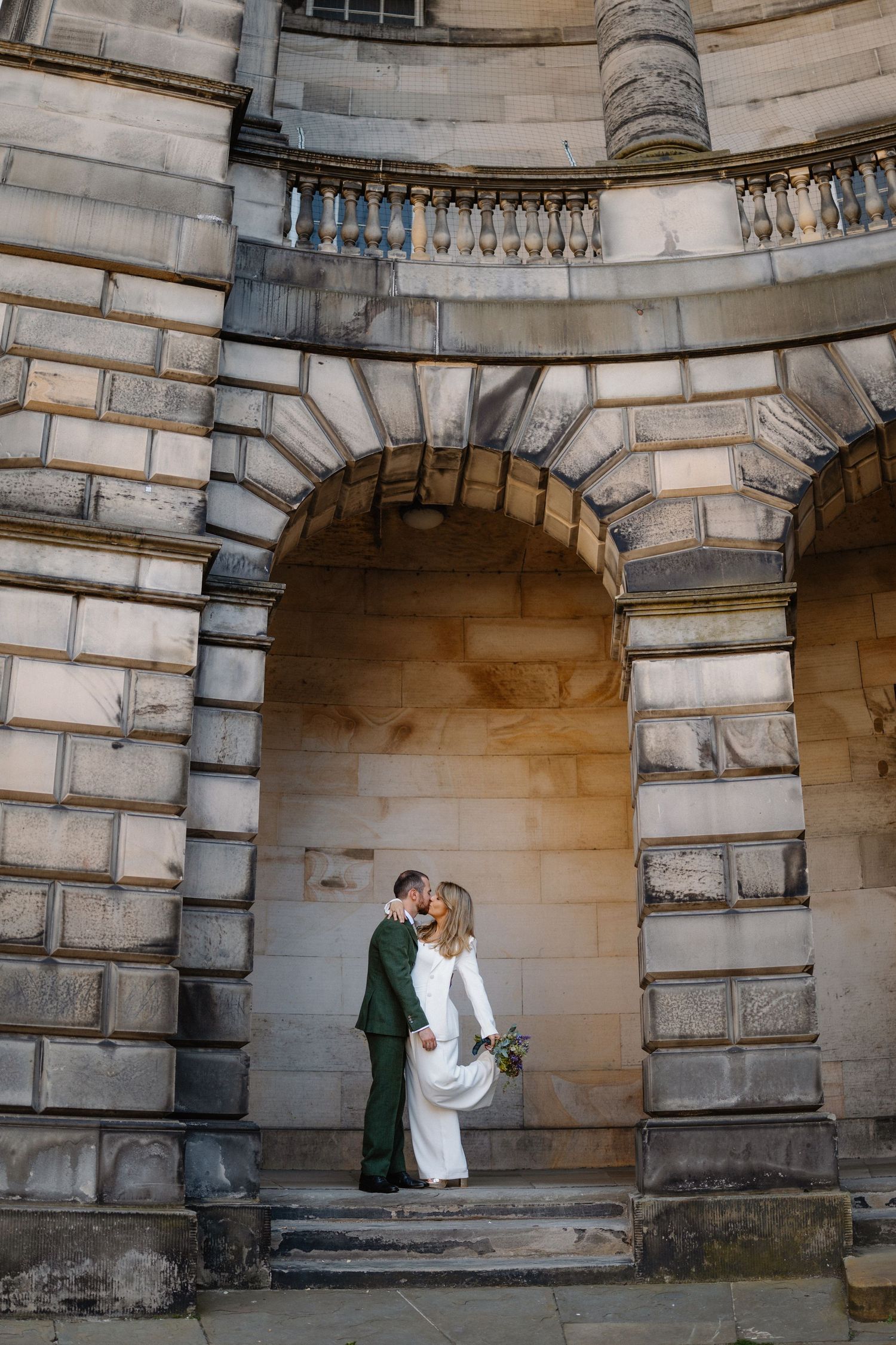 Bride and groom share a kiss under a grand stone archway at an elegant historic building during their wedding day.