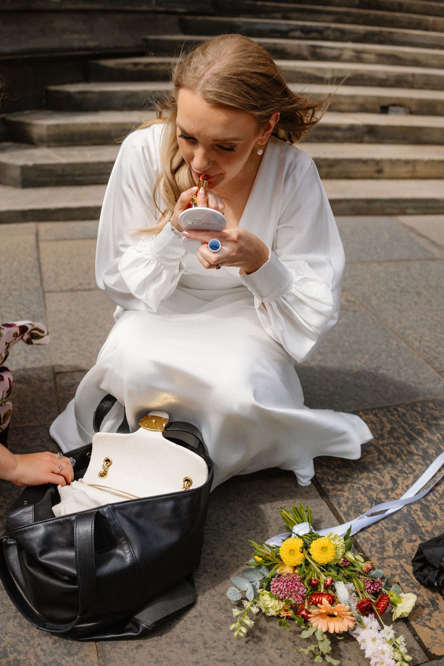 Bride in white satin gown touching up makeup while sitting on stone steps with bouquet nearby.