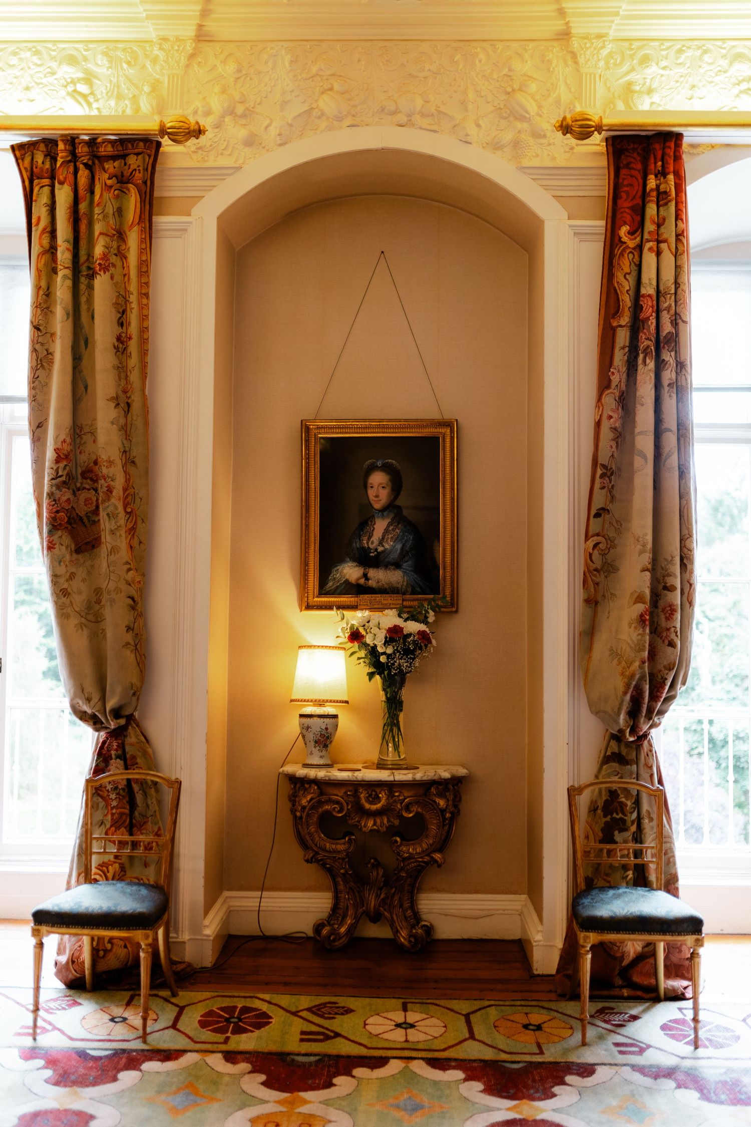 Elegant Georgian interior with ornate curtains, antique console table, floral arrangement, and portrait painting in arched alcove.